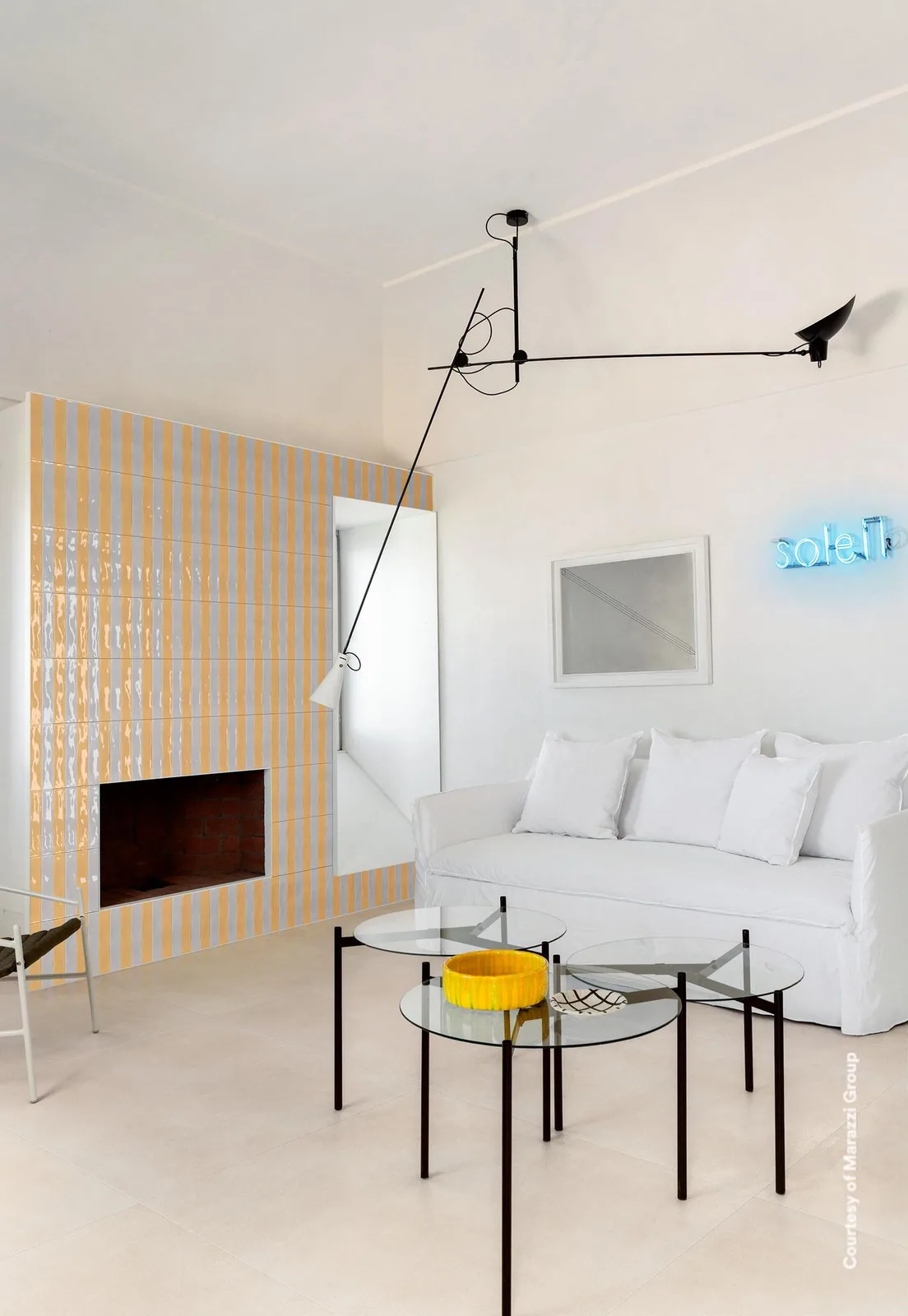 Kaminwand aus gelben und weißen Glanzriemchen in senkrechten Streifen, davor weißes Sofa und gläserne Beistelltische