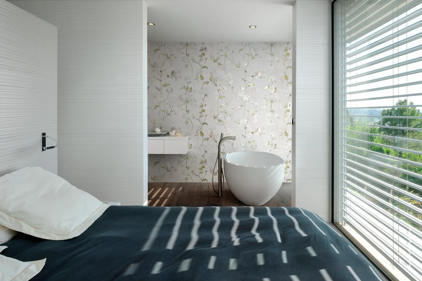Schlafzimmer mit weißer Rillenwand und Blick ins offene Bad mit Wanne vor einer Wand mit zarten Blütenranken.