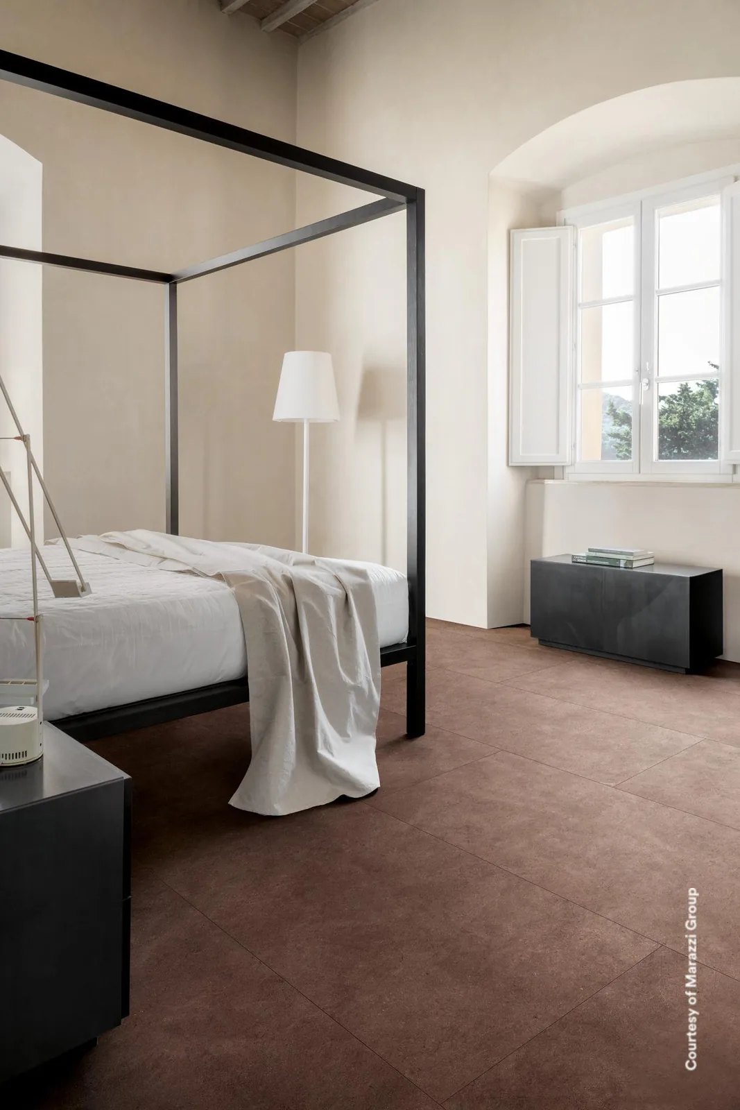 Schlafzimmer mit schwarzem Himmelbett, weißen Wänden und großformatigem dunkelbraunem Boden in Betonoptik.