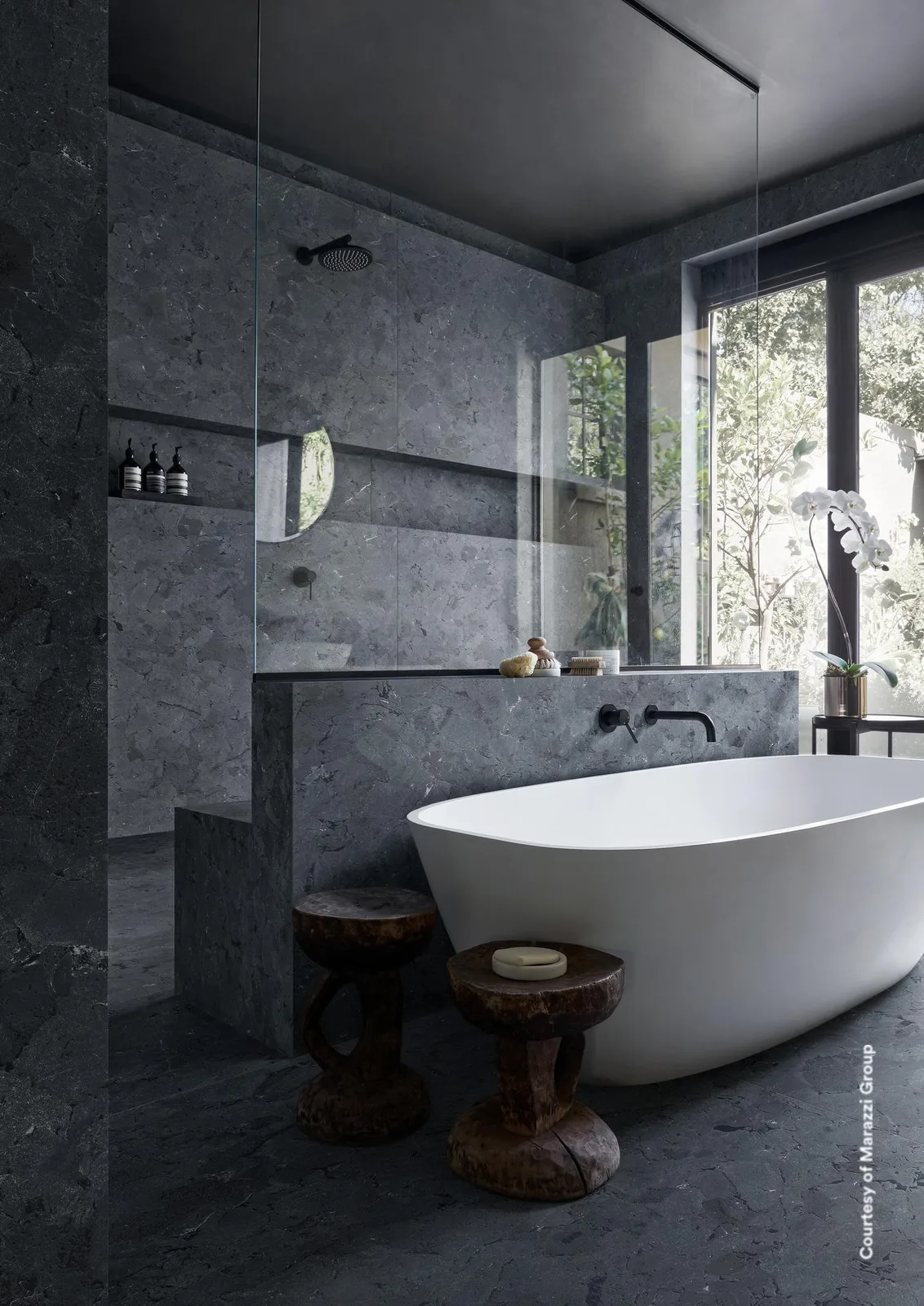 Bad mit Wänden und Boden in anthrazitfarbener Steinoptik, freistehender weißer Wanne und Holzhockern.