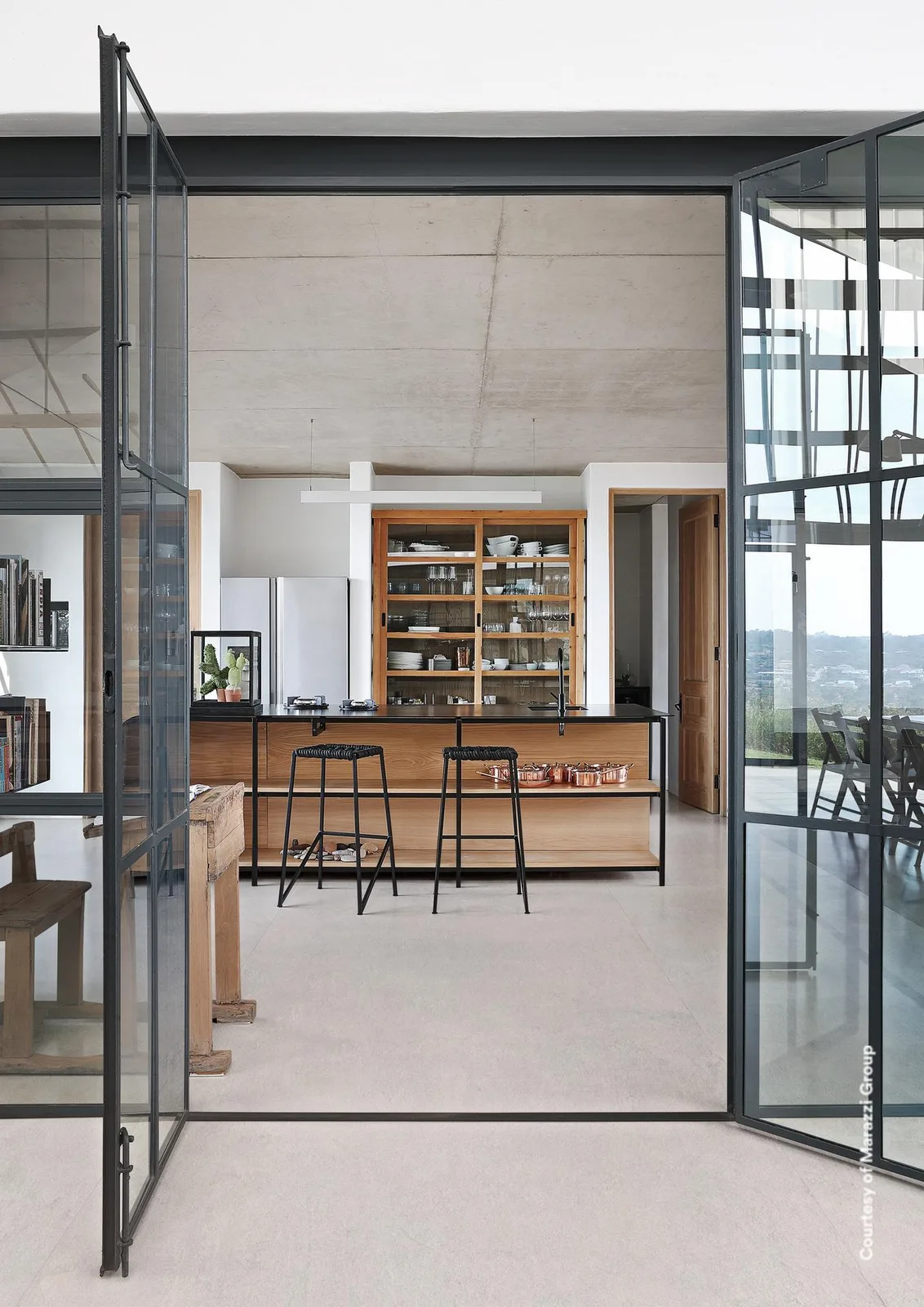 Blick durch eine schwarze Glastür in eine Küche mit Holzregalen und Barhockern, Boden in kalkweißer Betonoptik.