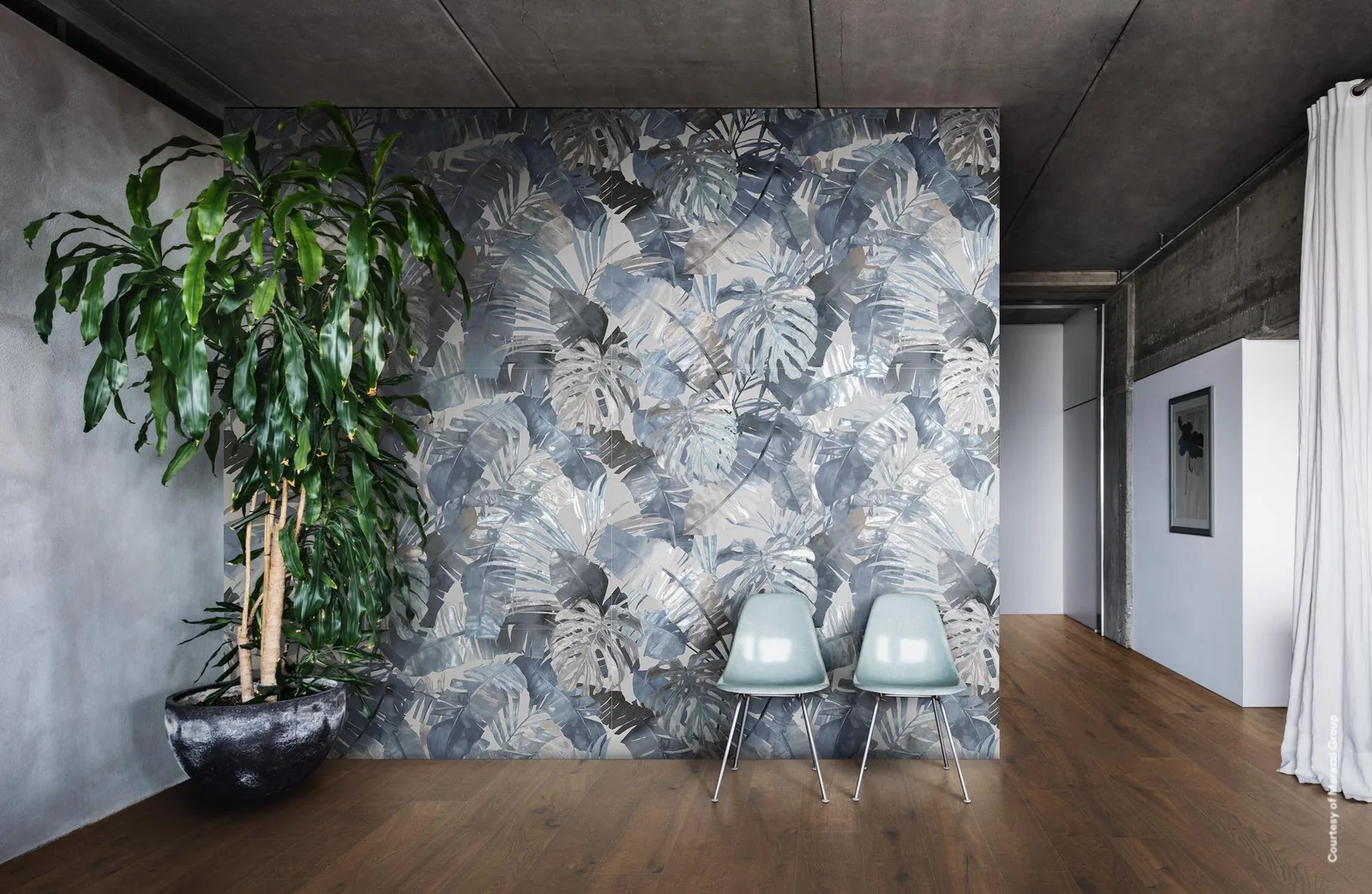 Raum mit Betondecke, Holzboden und zwei Stühlen vor einer Wand mit großen Tropenblättern in Blau und Grau, links eine Palme.