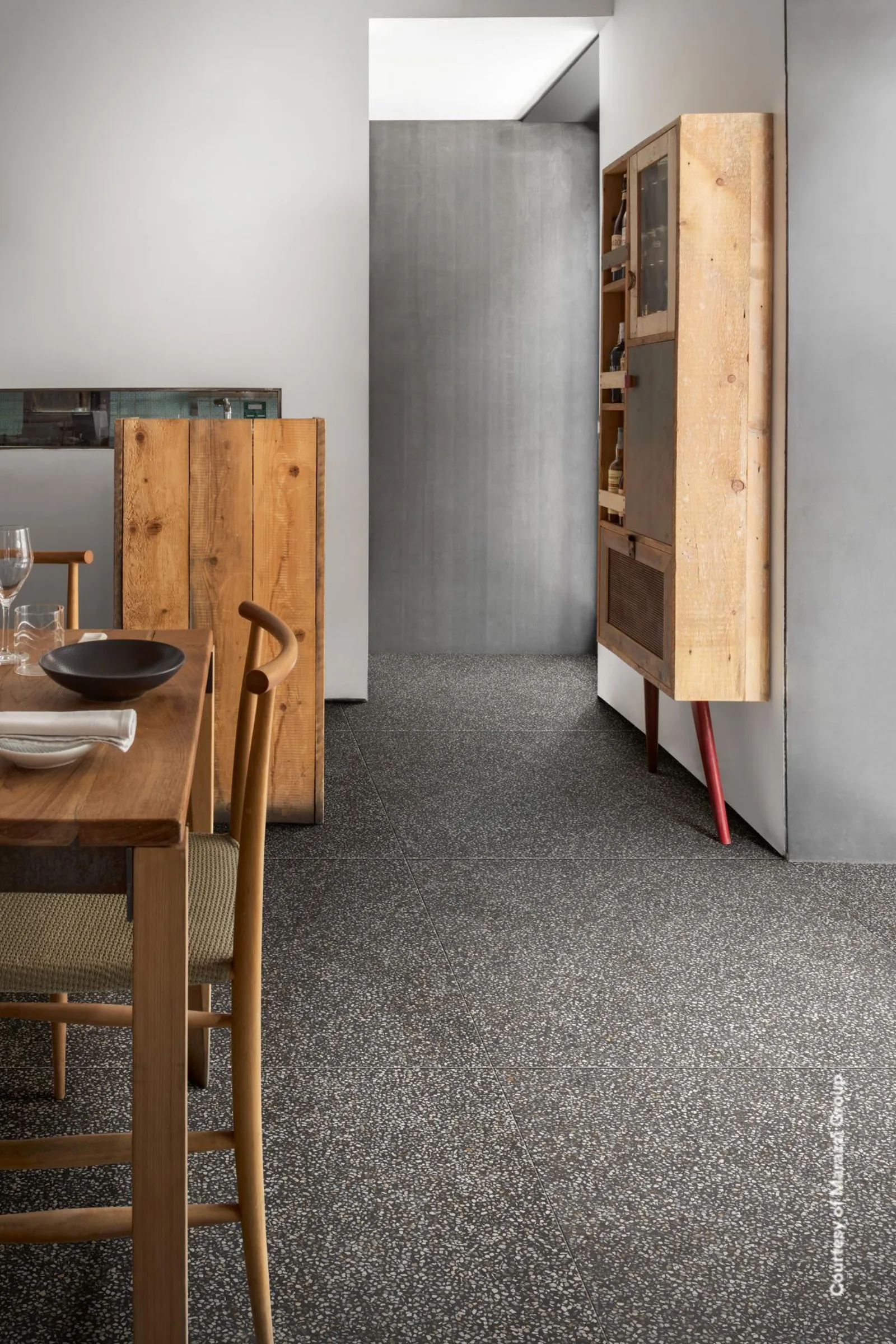 Essplatz mit Holzmöbeln vor einem offenen Durchgang, dunkler anthrazitfarbener Terrazzo-Boden mit hellen Splittern.