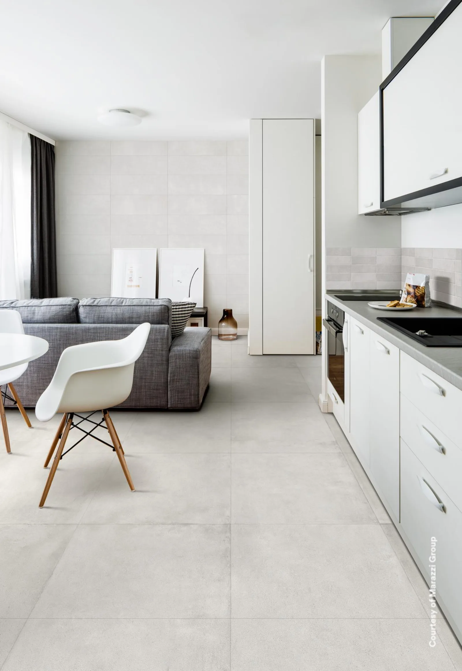 Wohnküche mit grauem Sofa, weißem Stuhl und heller Küchenzeile auf großformatigem Bodenbelag.