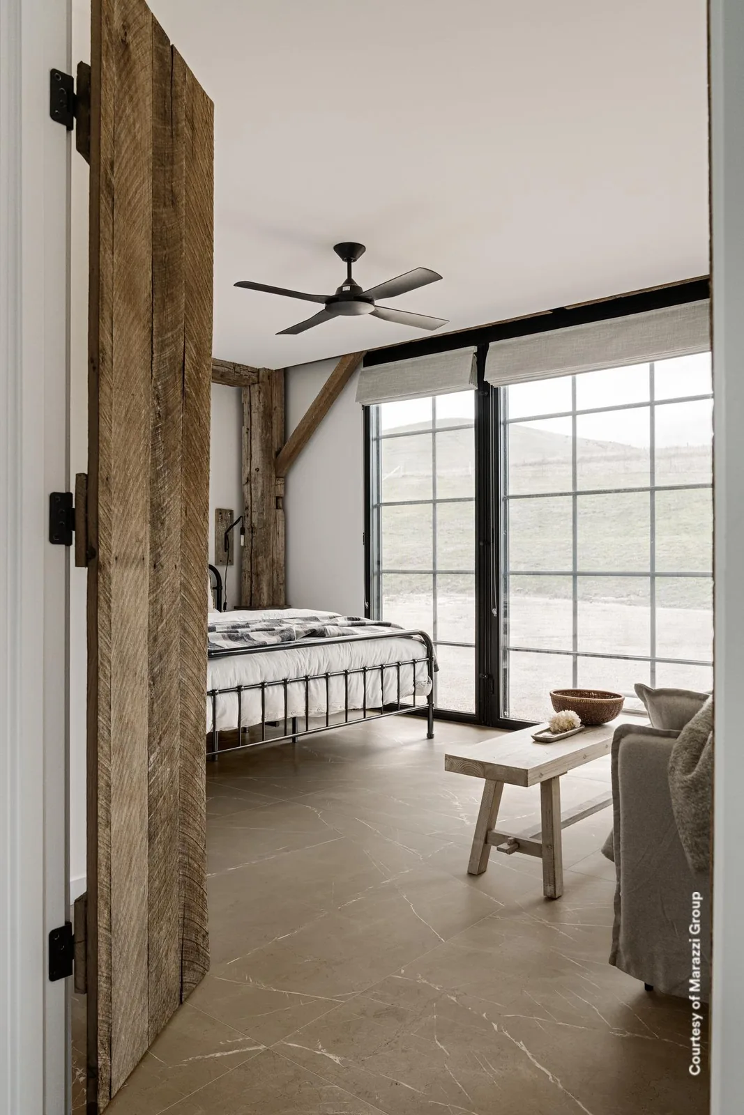Schlafzimmer mit Holztür, Metallbett und Fensterfront, Boden in graubeiger Marmoroptik mit feinen weißen Adern.
