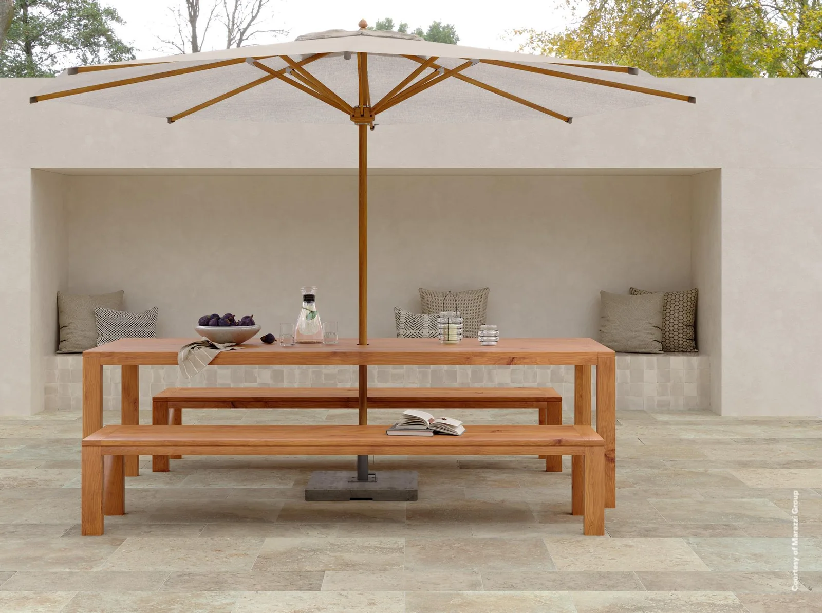 Terrasse mit Sonnenschirm, Holztisch und Bänken auf hellbeigen Steinoptik-Fliesen vor einer weißen Mauer