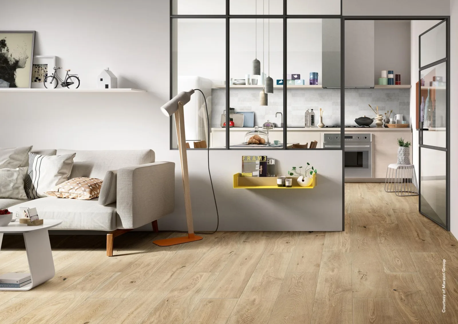 Offener Wohnraum mit hellem Holzoptik-Boden, grauem Sofa, Stehlampe und schwarz gerahmter Glaswand zur Küche.