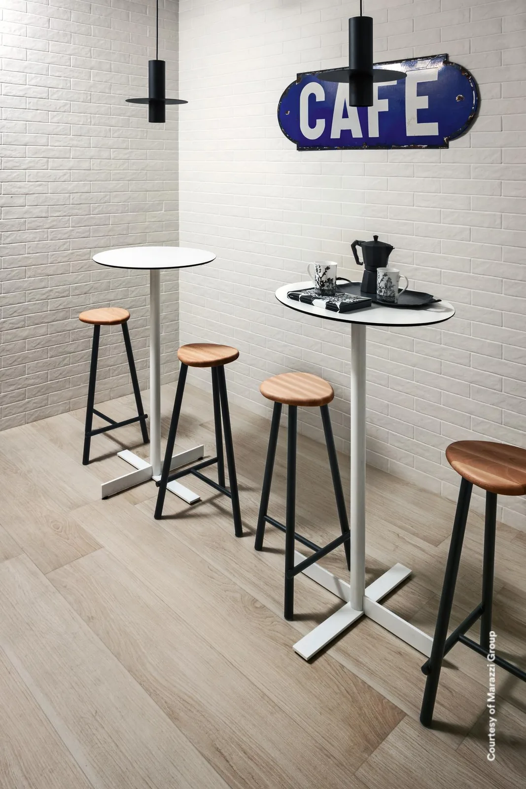 Bistroecke mit hellem Holzoptik-Boden, zwei weißen Stehtischen, vier Barhockern und weißer Ziegelwand mit Schild.