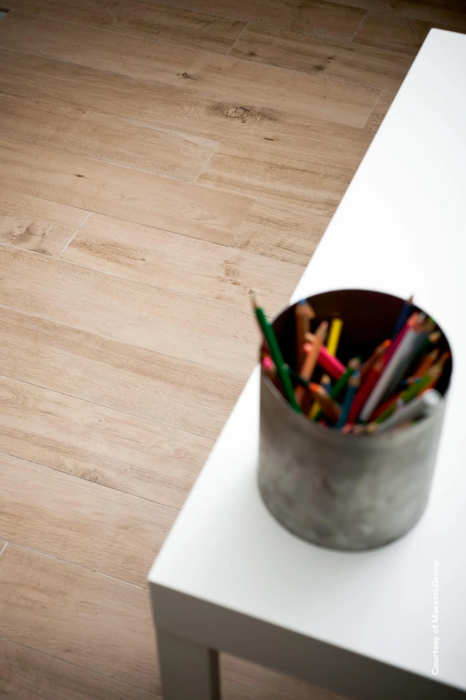 Dielenboden in warmer Holzoptik mit Astlöchern, darüber die Ecke eines weißen Tisches mit einem Becher Buntstifte.
