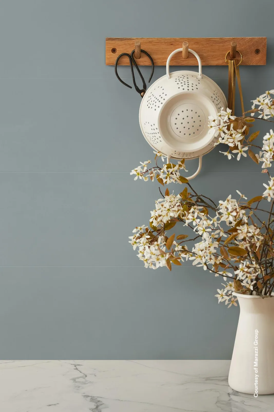 Küchenwand mit graublauen matten Wandfliesen, Hakenleiste mit Sieb und Vase mit weißen Blütenzweigen auf Marmor.