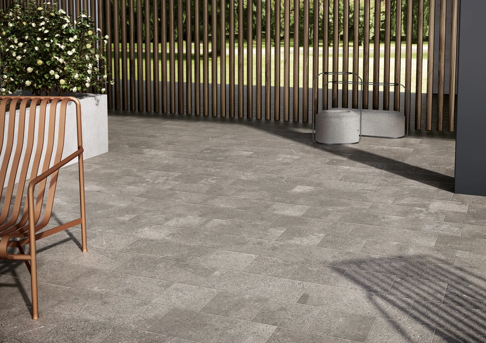 Terrasse mit grauen Steinoptik-Fliesen in gemischten Kleinformaten, Holzlamellenwand, Korbsessel und Pflanzkübel.