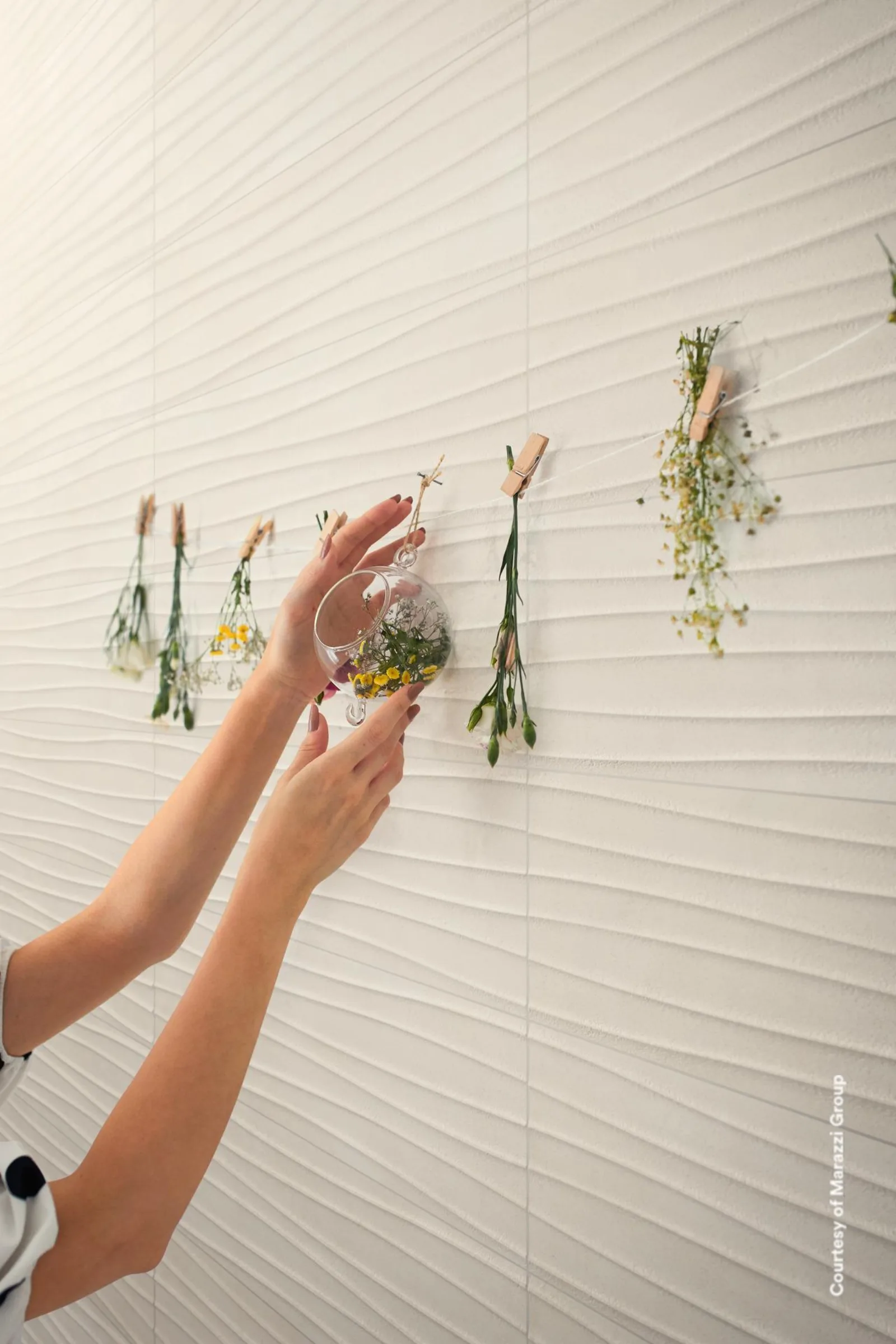 Zwei Hände legen kleine Trockenblumen an eine cremefarbene Wandfläche mit welligem Reliefmuster.