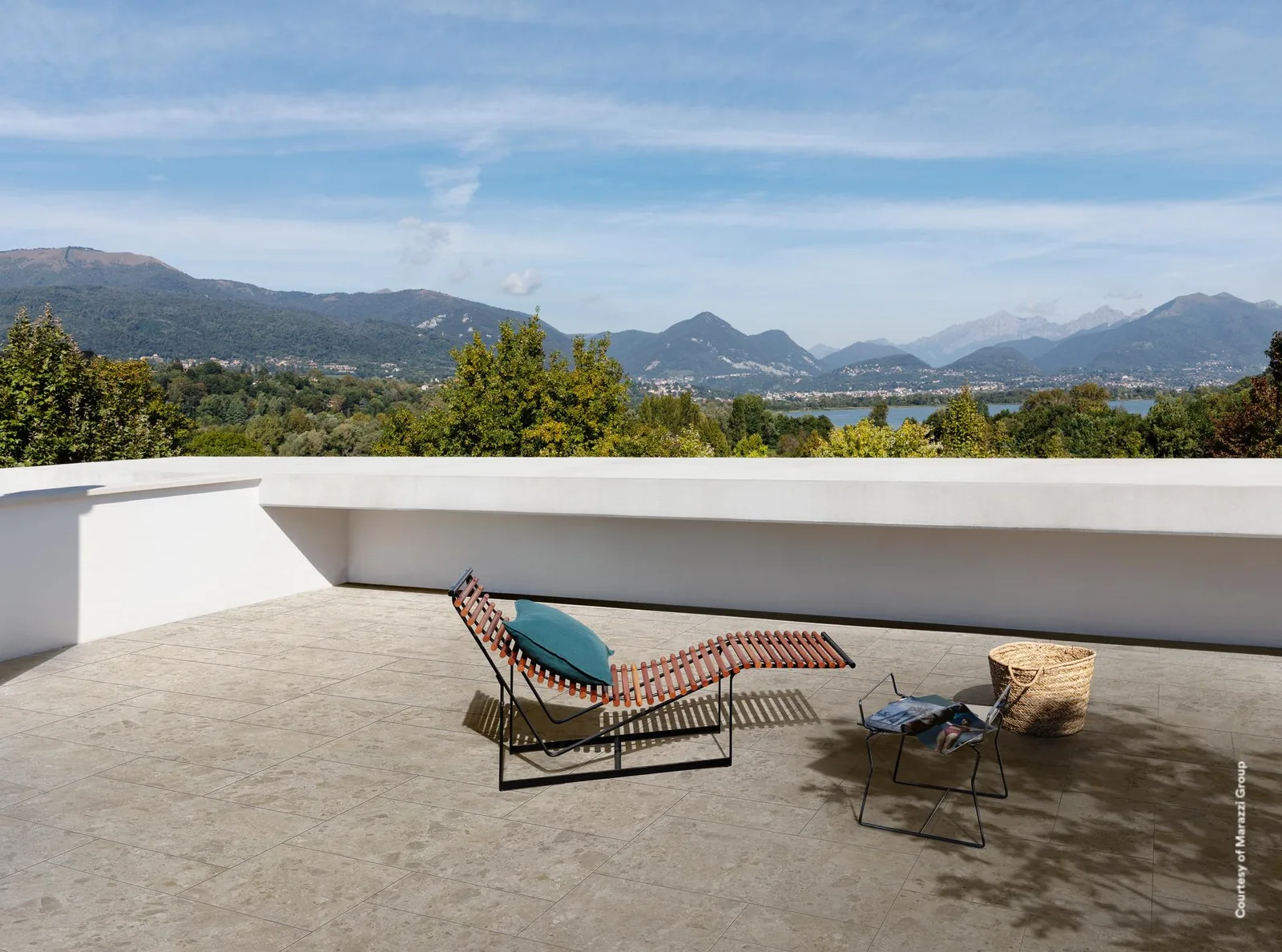 Dachterrasse mit Boden in beiger Terrazzo-Optik, zwei Liegen und Beistelltisch, dahinter Bergpanorama und See.