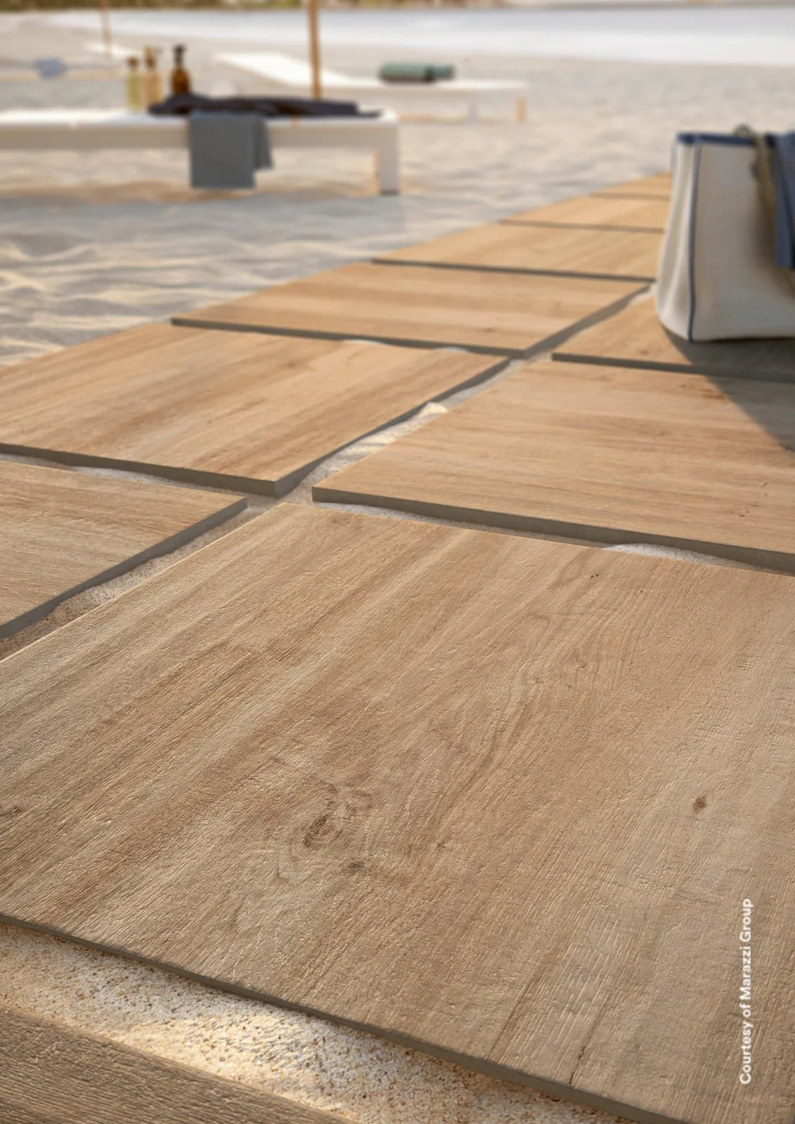 Marazzi Treverkhome20 Rovere: Nahaufnahme verlegter Holzoptik-Dielen im Sand, Liege und Strandtasche im Hintergrund