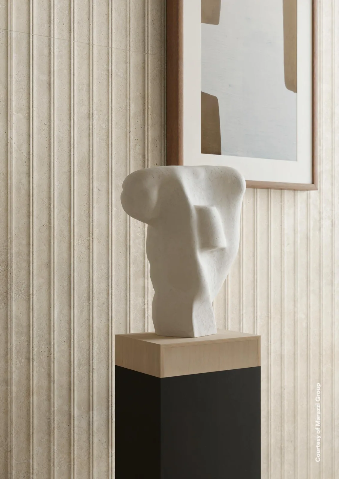 Wand aus beigen Rillenfliesen in Travertinoptik mit Skulptur auf einem Sockel und gerahmtem Bild davor