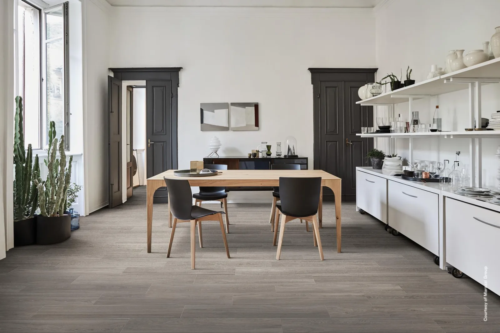 Essbereich mit grauem Holzoptik-Boden, Holztisch mit schwarzen Stühlen, weißen Regalen und Kakteen am Fenster.