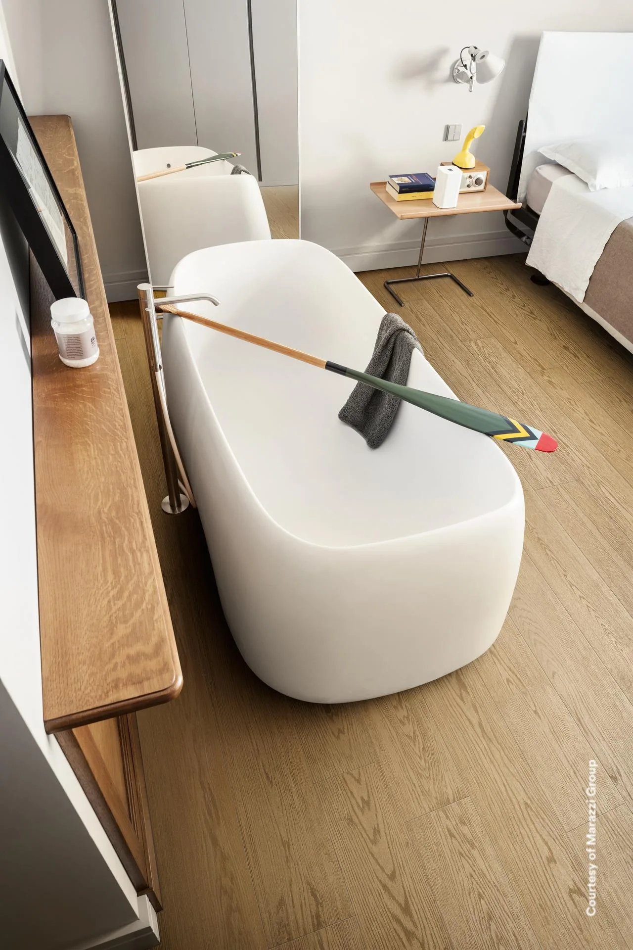 Bad mit freistehender ovaler Wanne und Holzkonsole auf hellen Dielen in Holzoptik, Bett im Hintergrund.