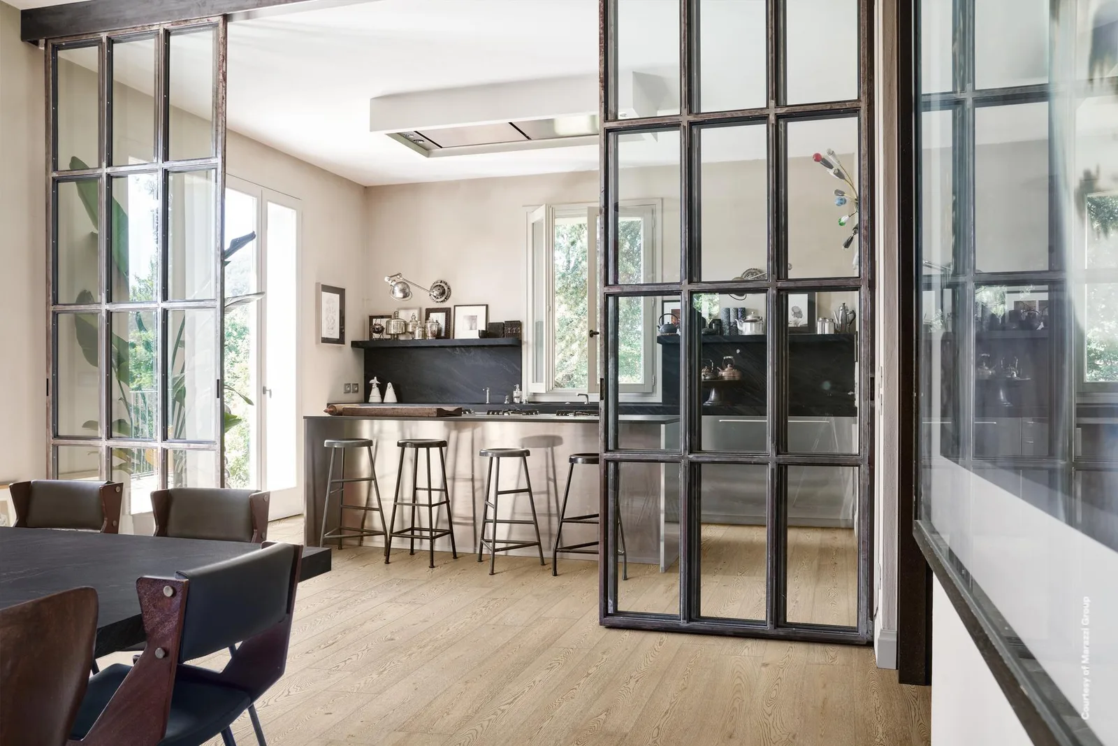 Offene Küche mit Theke und Barhockern hinter einer Stahl-Glas-Wand, Boden in hellbeigen Holzoptik-Planken.