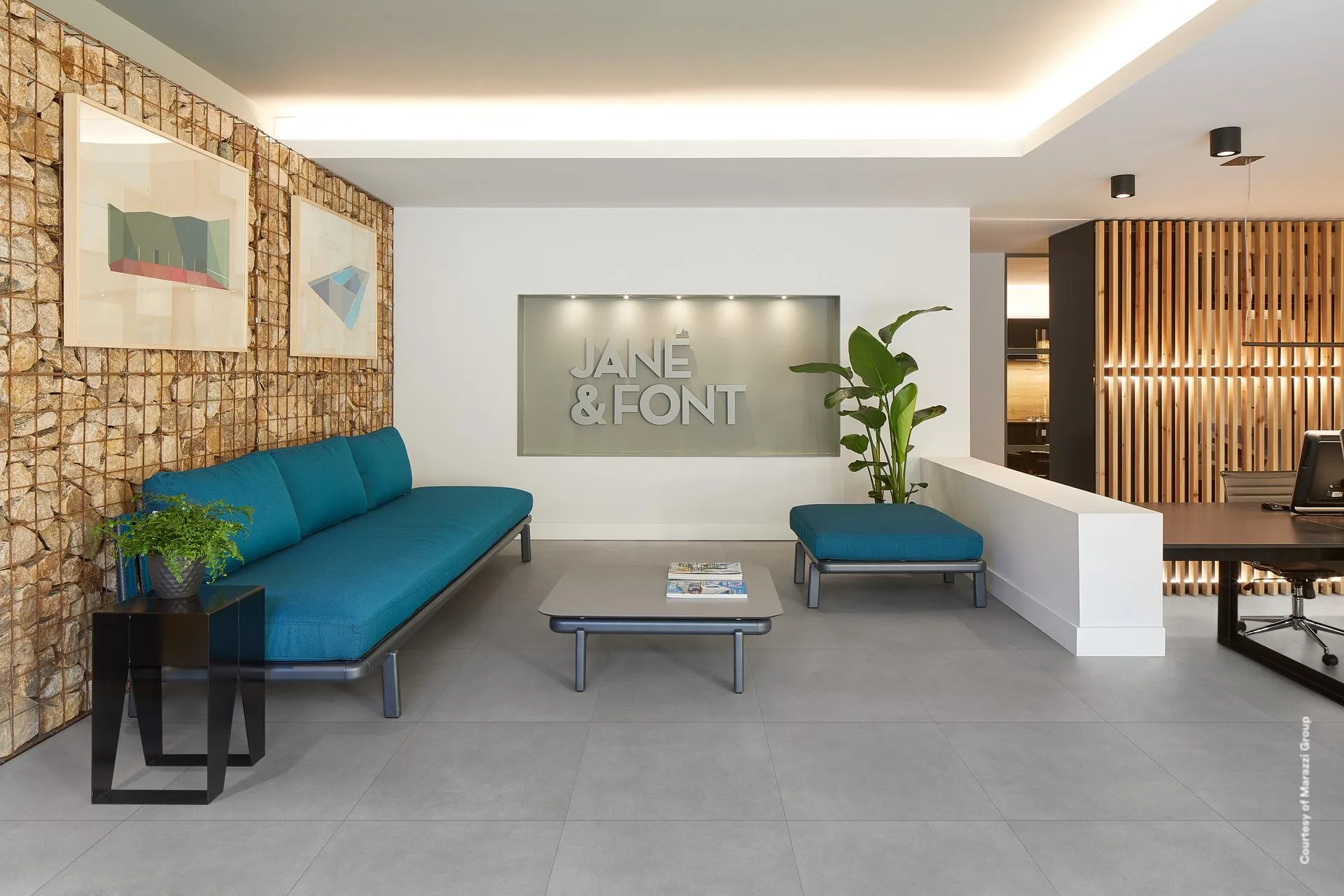 Büro-Lounge mit blauem Sofa, Natursteinwand und Holzlamellen, mittelgrauer Betonoptik-Boden im Format 60x60 cm.