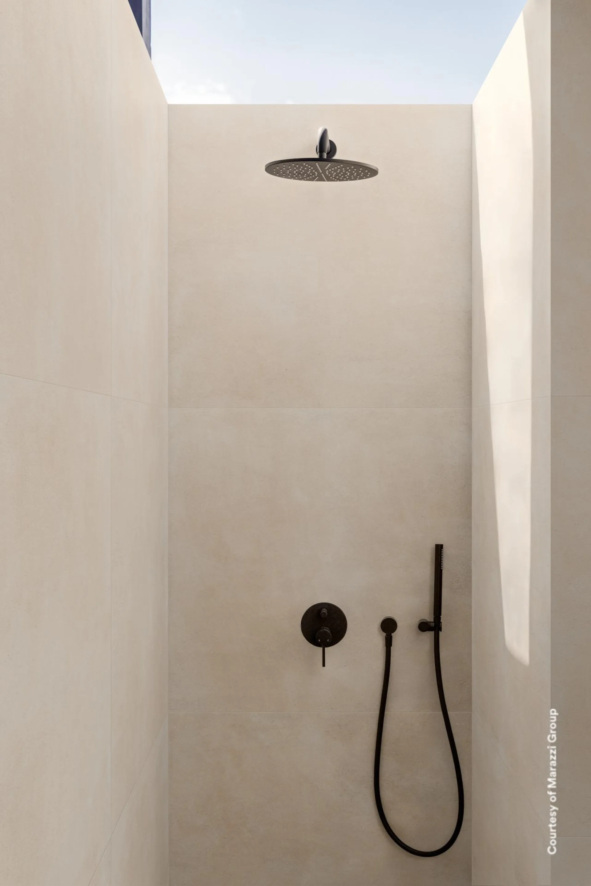 Offene Dusche mit hellbeigen Großformatfliesen an Wand und Boden, schwarzer Armatur und Tageslicht von oben.