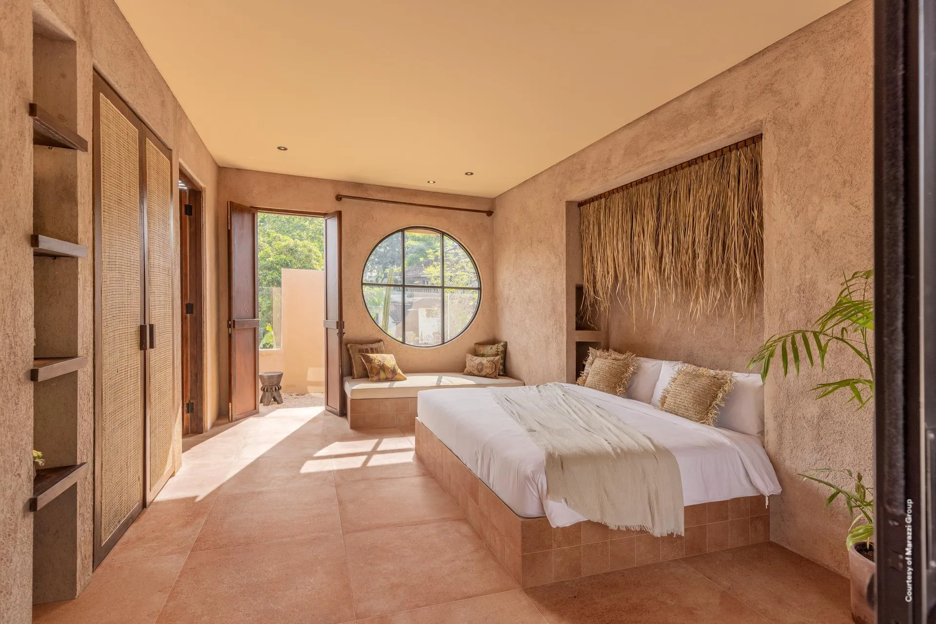 Schlafzimmer mit Bett, Bastwandbehang, Rundfenster und terrakottafarbenen Wänden auf rotbraunem Boden.