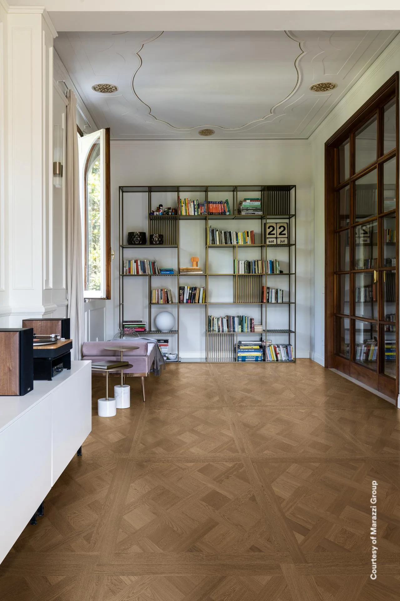 Bibliothek mit offenem Regal und Stuckdecke über dunkelbraunem Kassettenparkett in Holzoptik.