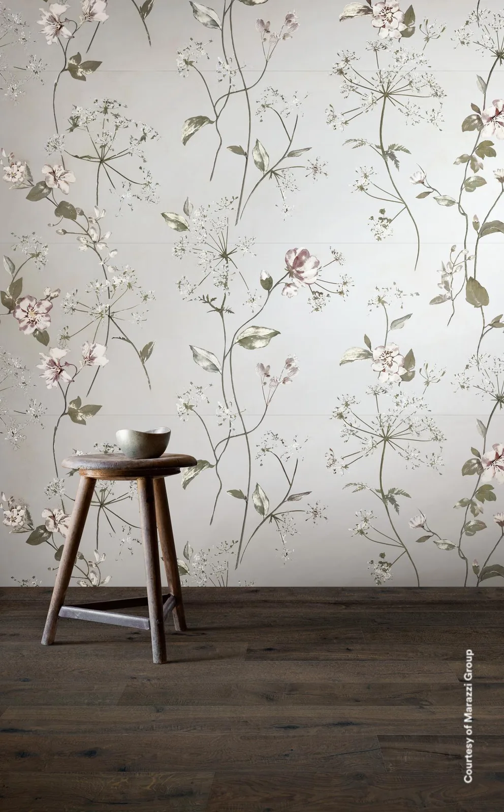 Hocker auf dunklem Holzboden vor einer hellen Wand mit feinen Gräsern und kleinen Blüten auf weißem Grund.