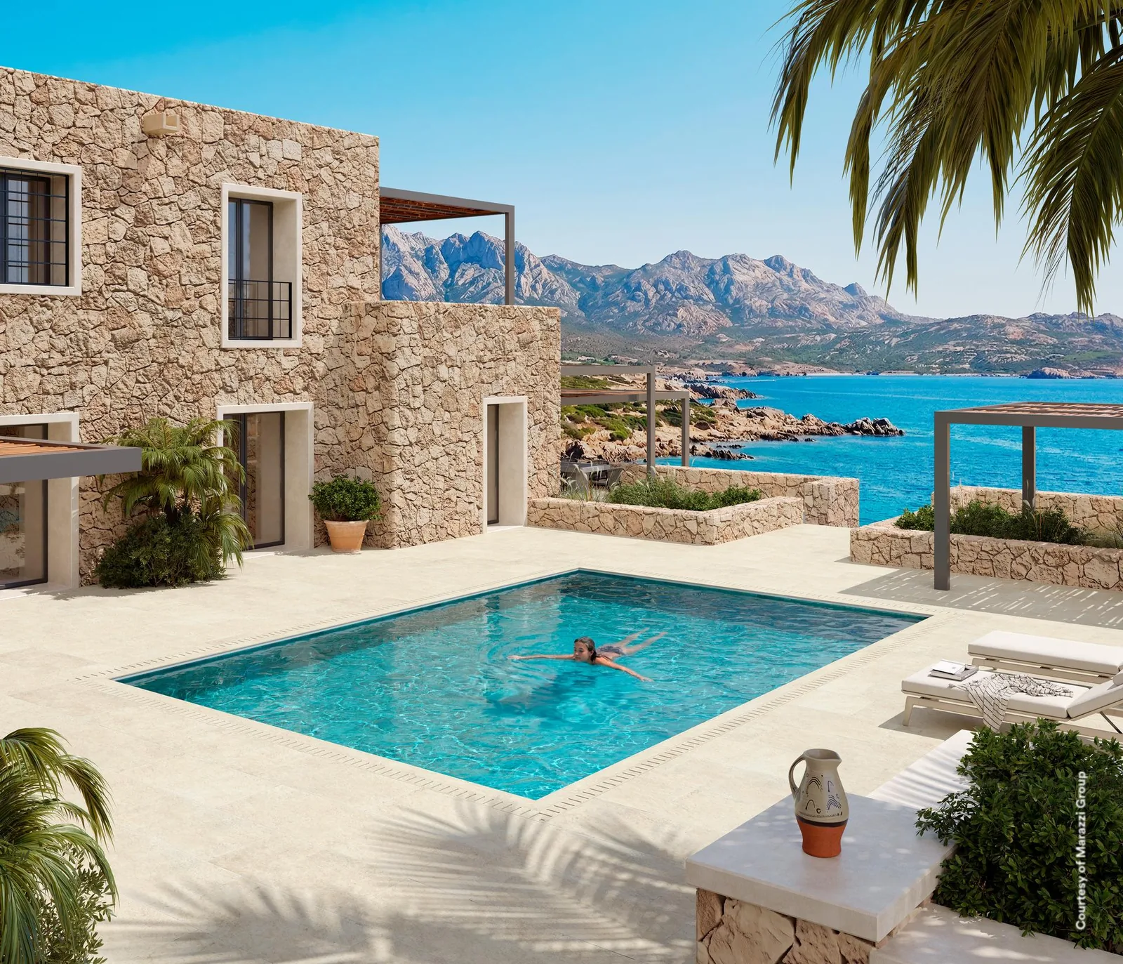 Pooldeck mit cremeweißen Großformatplatten vor einem Natursteinhaus, Sonnenliegen und Blick auf Meer und Berge.