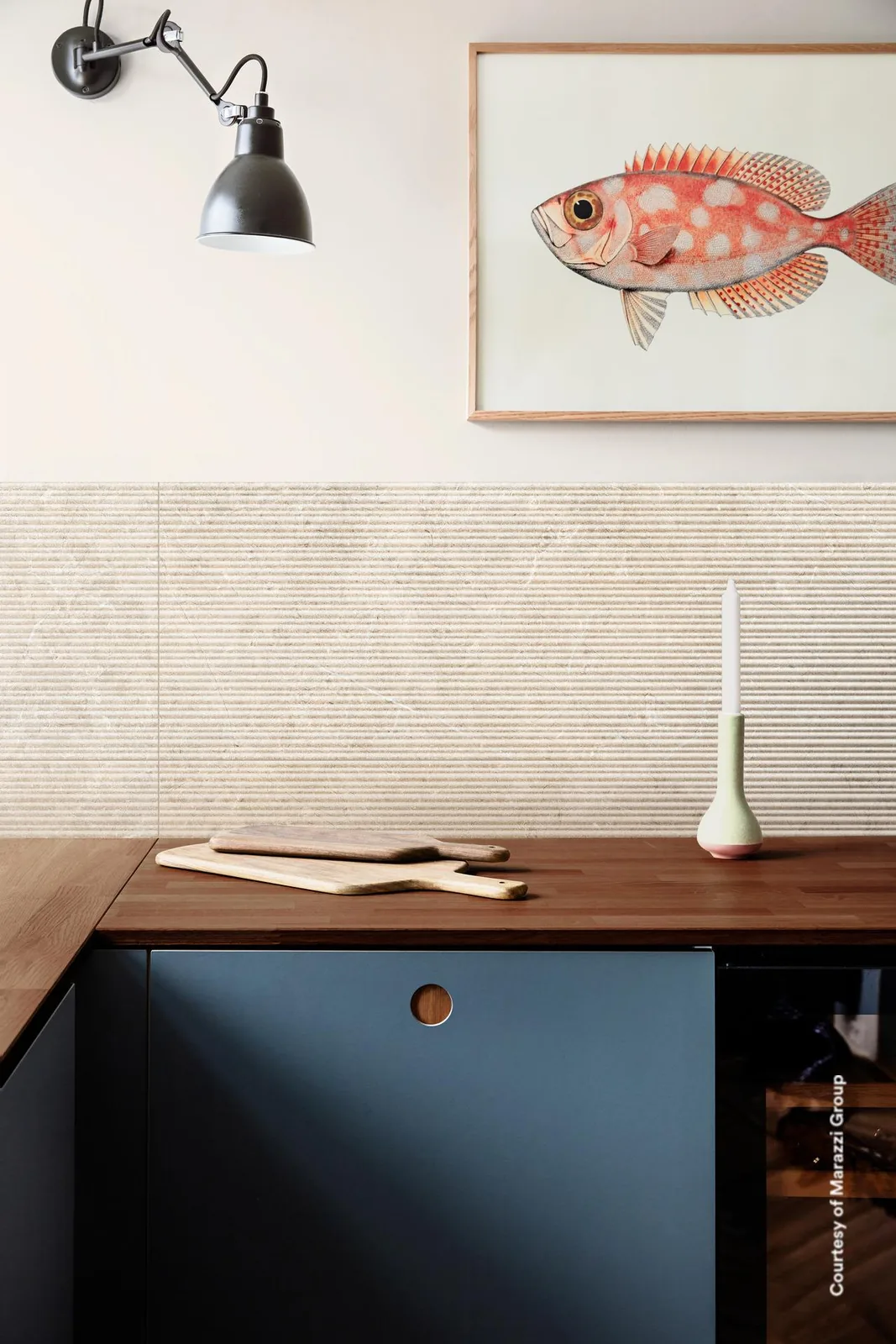 Küchenwand mit sandbeigen Fliesen mit Rillenrelief über einer Holzarbeitsplatte, darüber ein Fischbild und eine Leuchte.