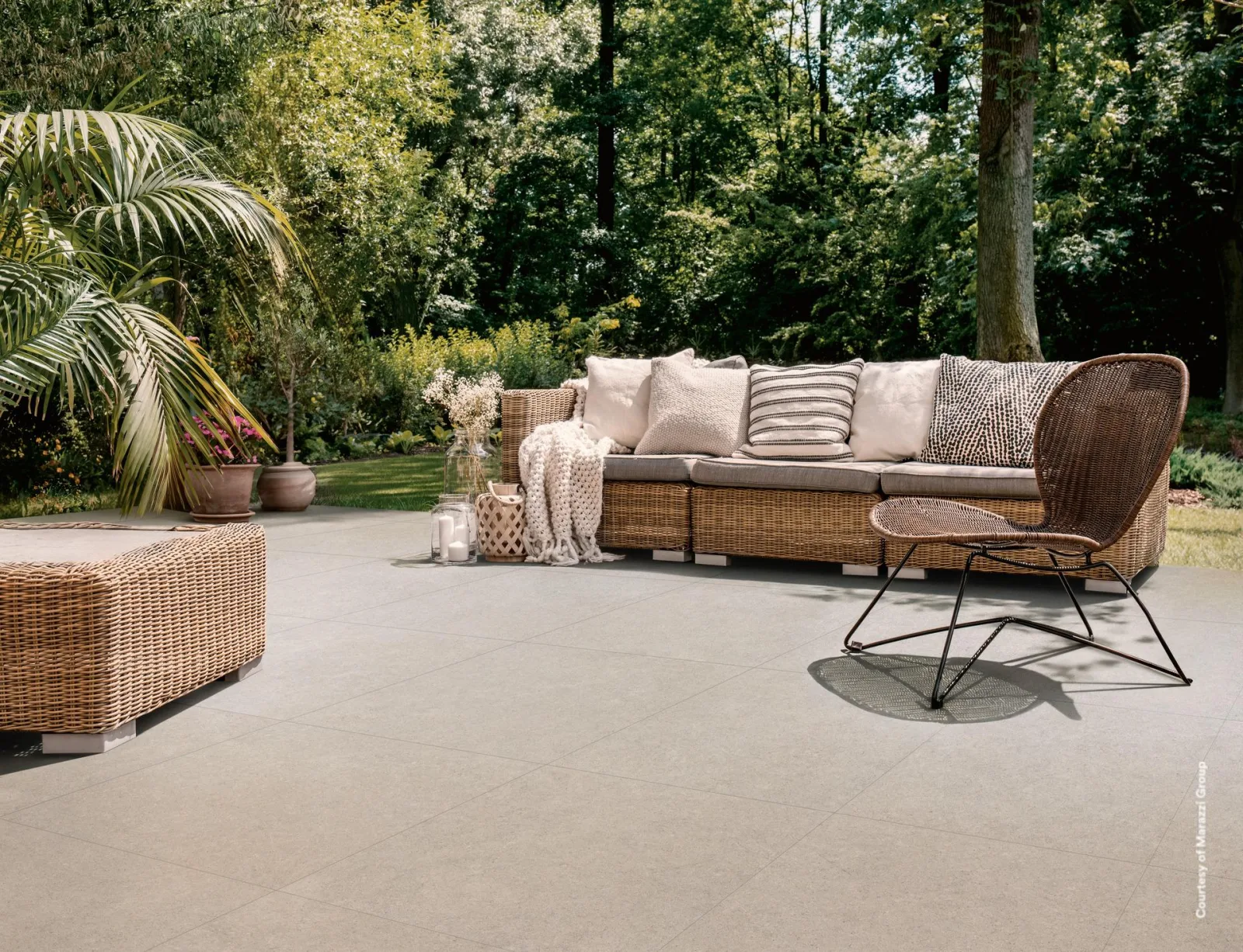 Terrasse mit sandfarbenen Steinoptik-Platten im Großformat, Rattan-Lounge mit Kissen, Palme und Bäume im Hintergrund.