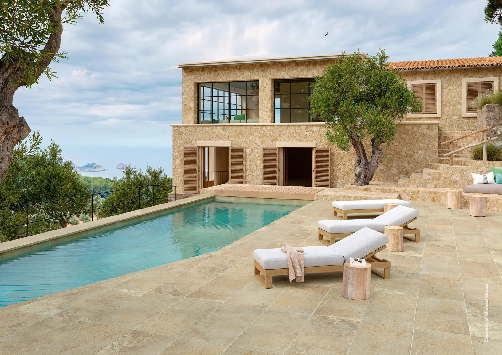 Poolterrasse mit warmbeigen Großformatplatten vor einem Steinhaus, drei Holzliegen und einem Olivenbaum.