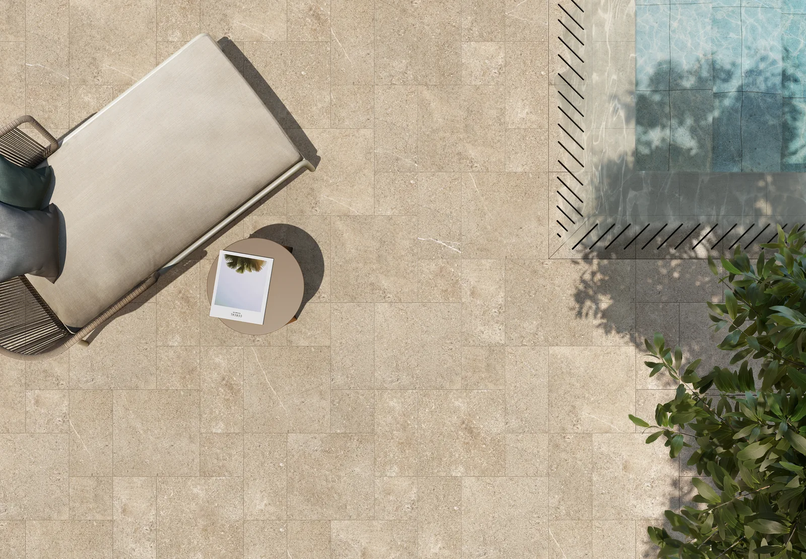 Poolterrasse von oben mit sandbeigen Steinoptik-Fliesen in gemischten Formaten, Liege und Beistelltisch am Beckenrand.