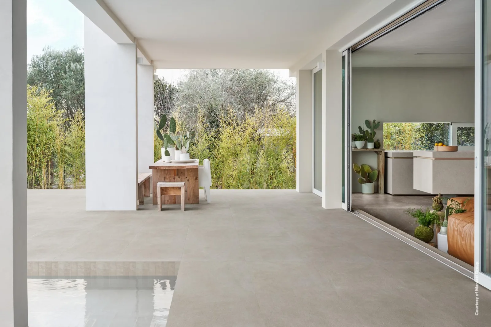 Überdachte Terrasse mit greige Betonoptik-Platten im Großformat, Poolrand, Holztisch mit Kakteen und Blick in den Garten.