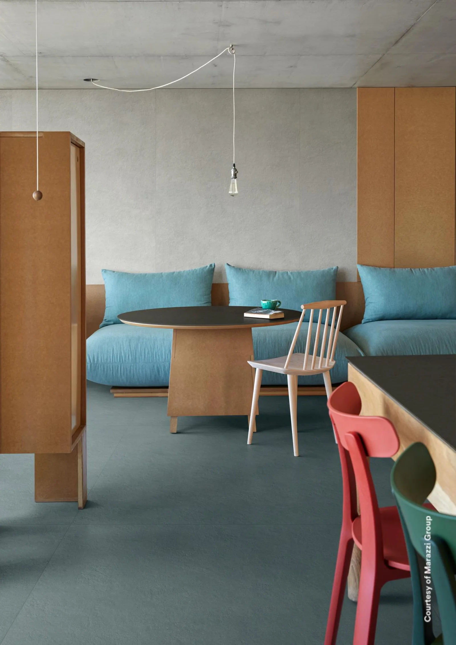 Wohnraum mit türkisfarbenem Sofa, rundem Tisch und Holzschränken auf blaugrauem Betonoptik-Boden im Großformat.