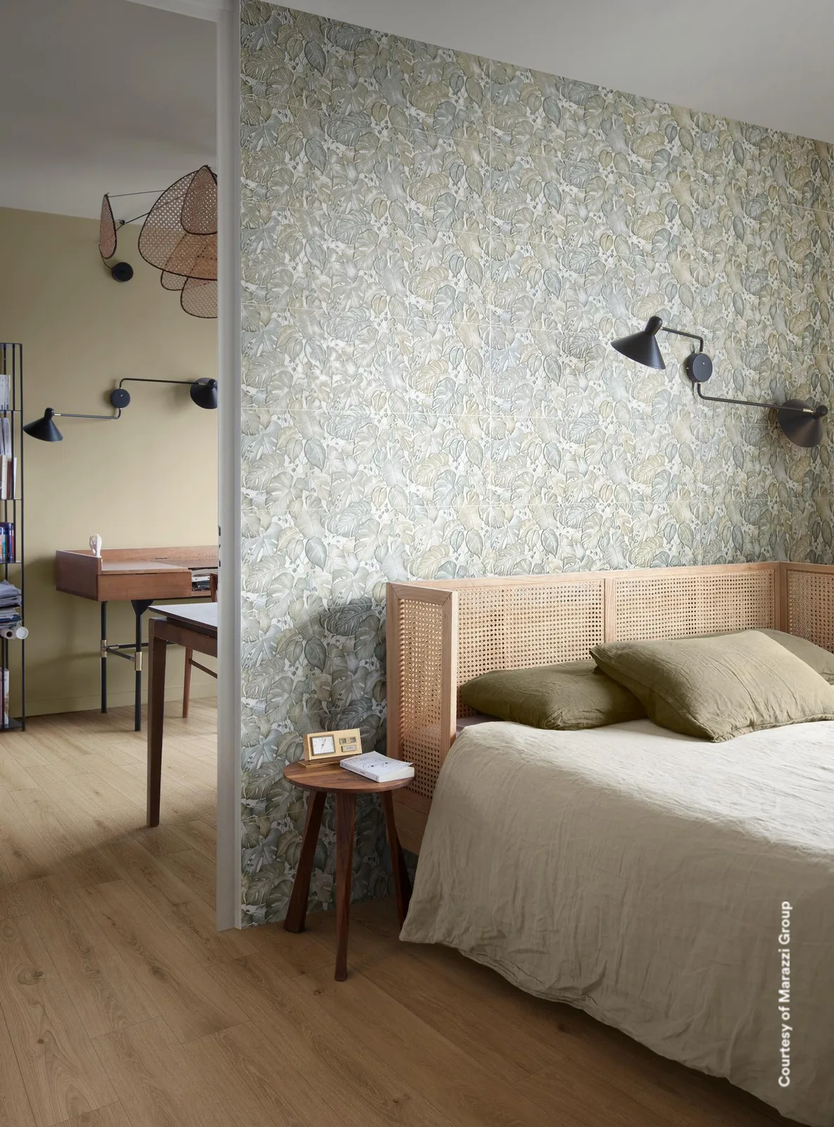 Schlafzimmer mit Bett vor einer Trennwand aus Fliesen mit graugrünem Blattmuster, daneben Schreibtisch und Wandleuchte.