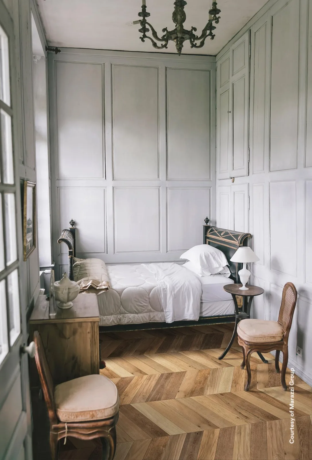 Schlafzimmer mit weißer Wandvertäfelung und antikem Bett, Boden aus braunen Holzoptik-Fliesen im Chevron-Muster.