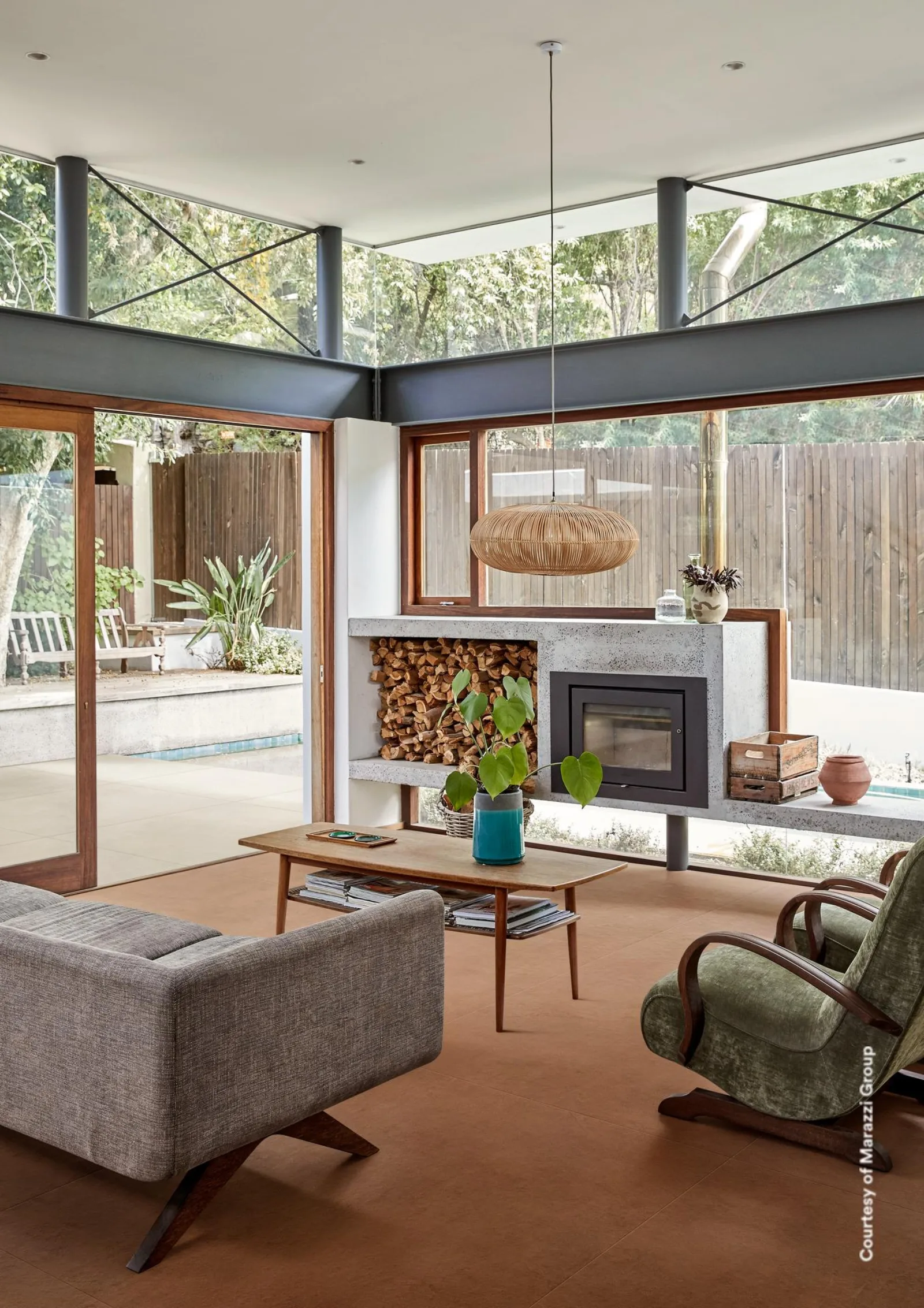 Wohnzimmer mit Kamin, grauem Sofa und Glasfront zum Garten auf terrakottabraunem Betonoptik-Boden.