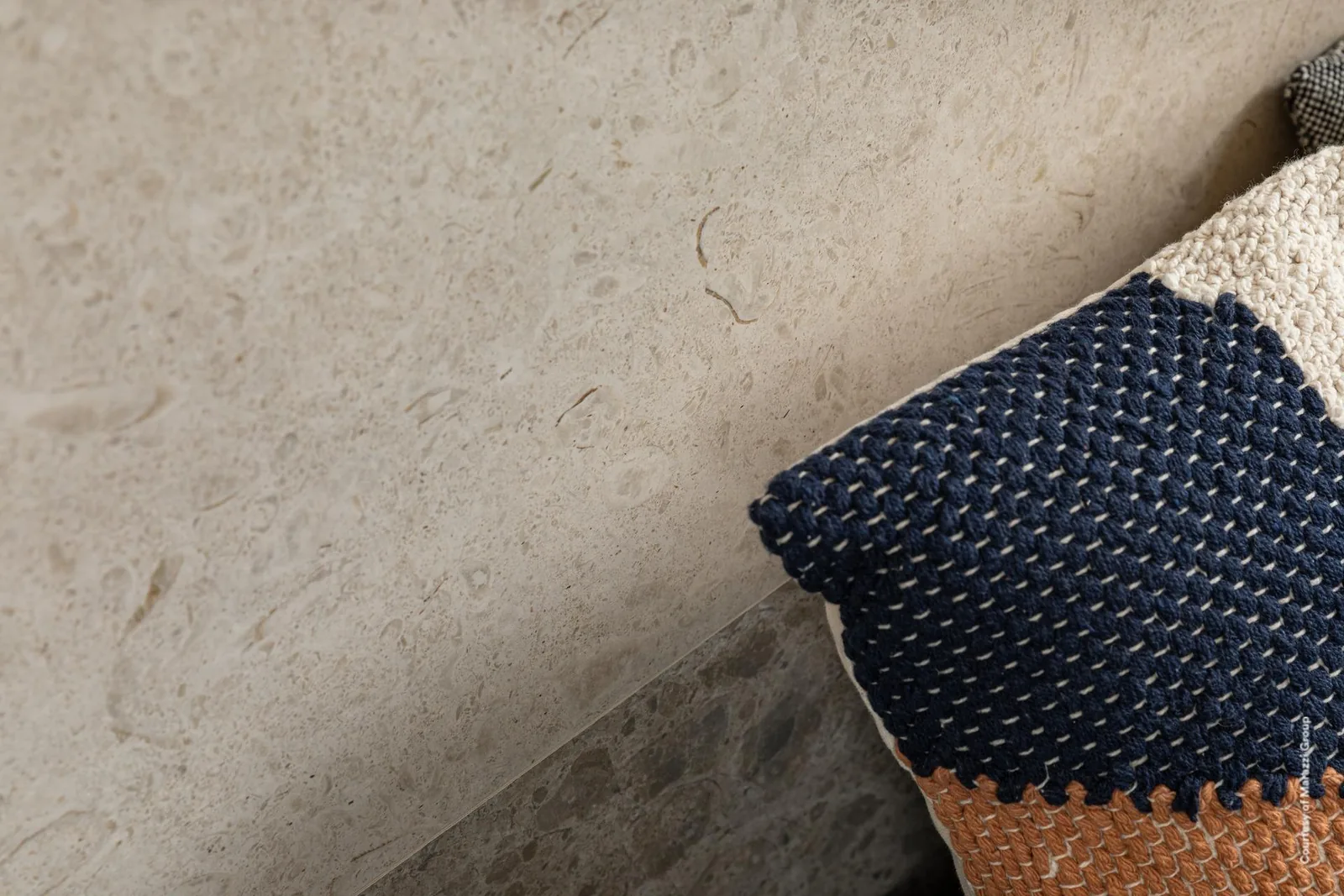 Nahaufnahme von sandbeiger Steinoptik-Wand und Terrazzo-Optik-Boden, darauf ein gemustertes Kissen in Blau und Orange.