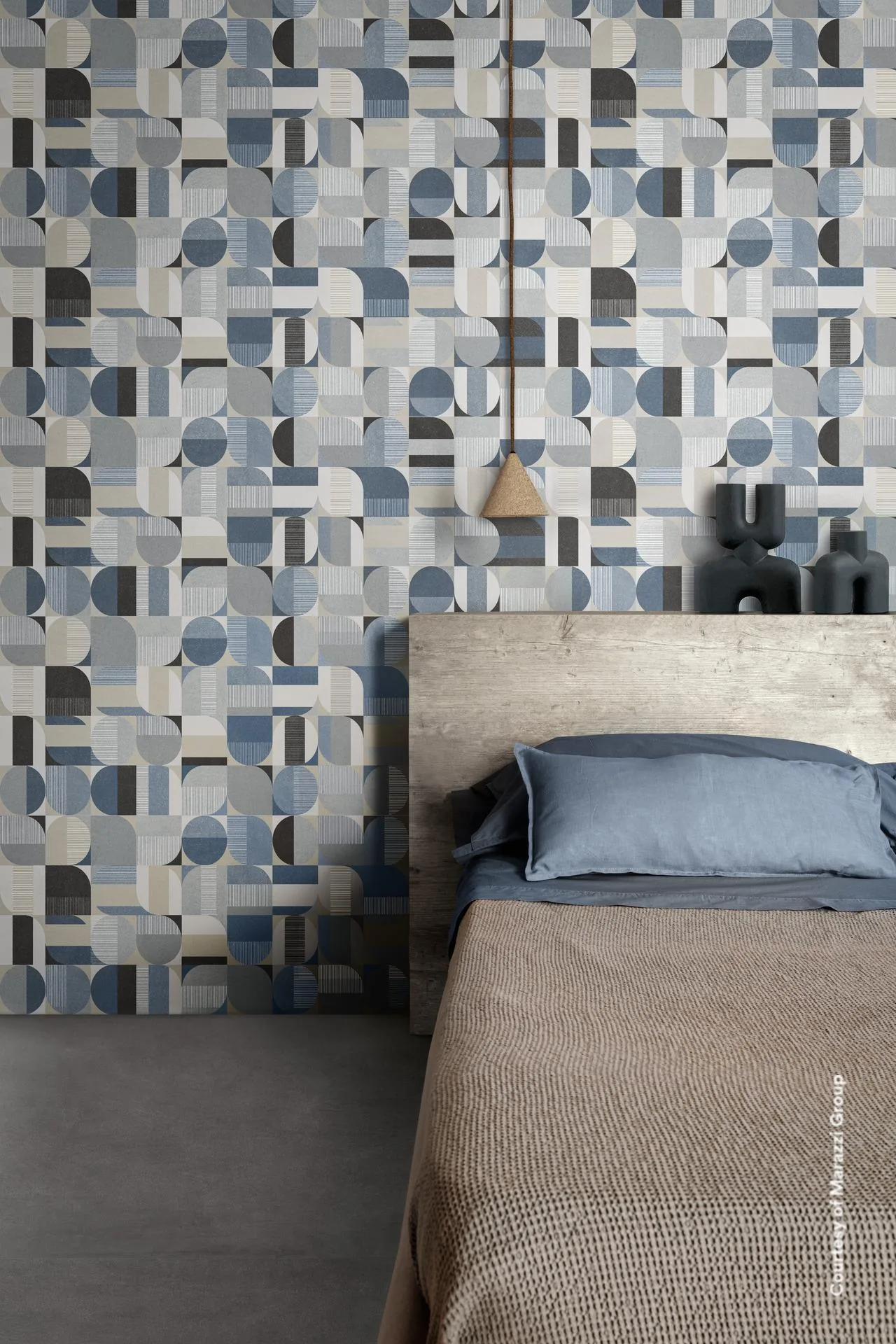 Schlafzimmer mit blau-grau gemusterter Wand aus Halbkreisen und Quadraten, Holzbett mit blauem Kissen und dunklem Boden