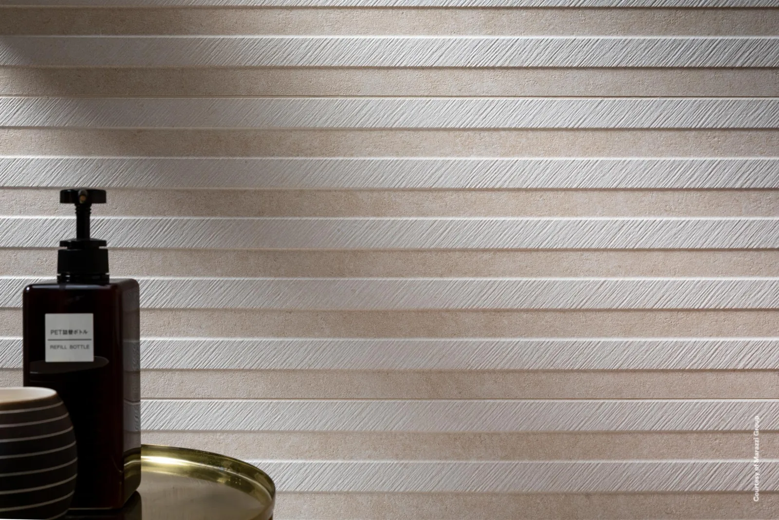 Wand aus sandbeigen Fliesen mit horizontalen Reliefstreifen, davor Seifenspender und Schalen auf goldenem Tablett.