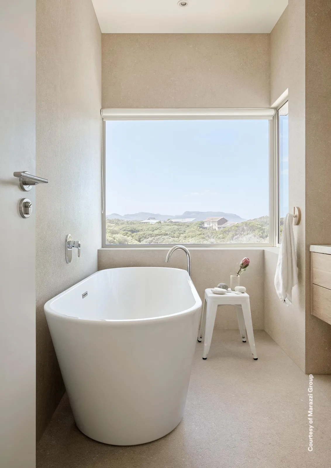 Bad mit Wänden und Boden in hellbeiger Steinoptik, freistehender Badewanne und großem Fenster.