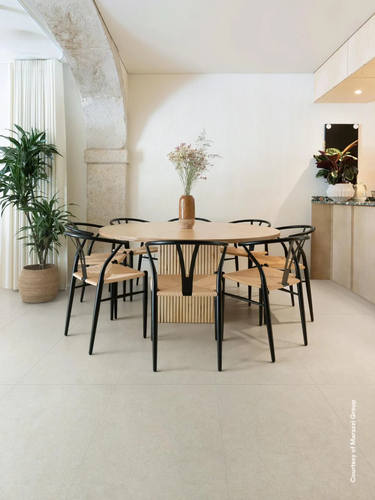 Essbereich mit cremeweißer Steinoptik-Fliese im Großformat, runder Holztisch mit Stühlen, Steinbogen und Pflanze.