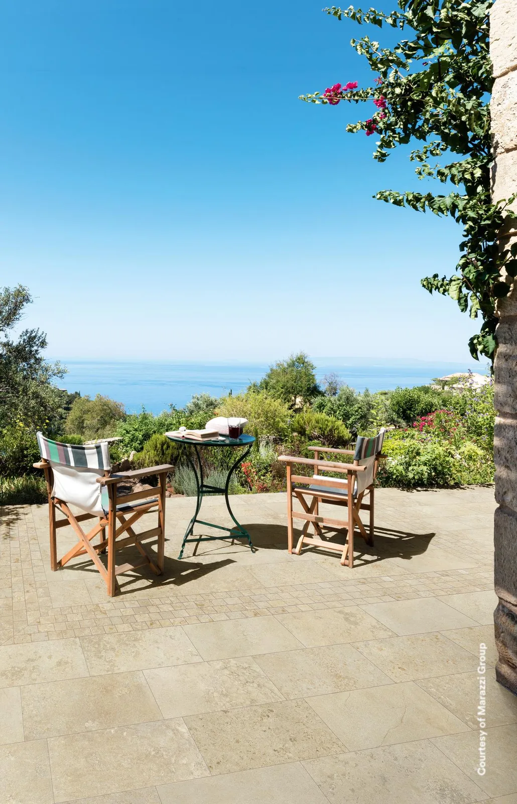 Gartenterrasse mit beigen Steinoptik-Bodenfliesen, zwei Holzstühle und Bistrotisch vor Sträuchern und Meerblick
