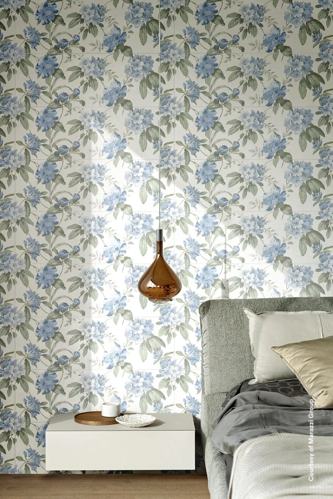 Schlafzimmer mit Bett und Pendelleuchte vor einer Wand aus weißen Fliesen mit großen blauen Blüten und grünen Blättern.