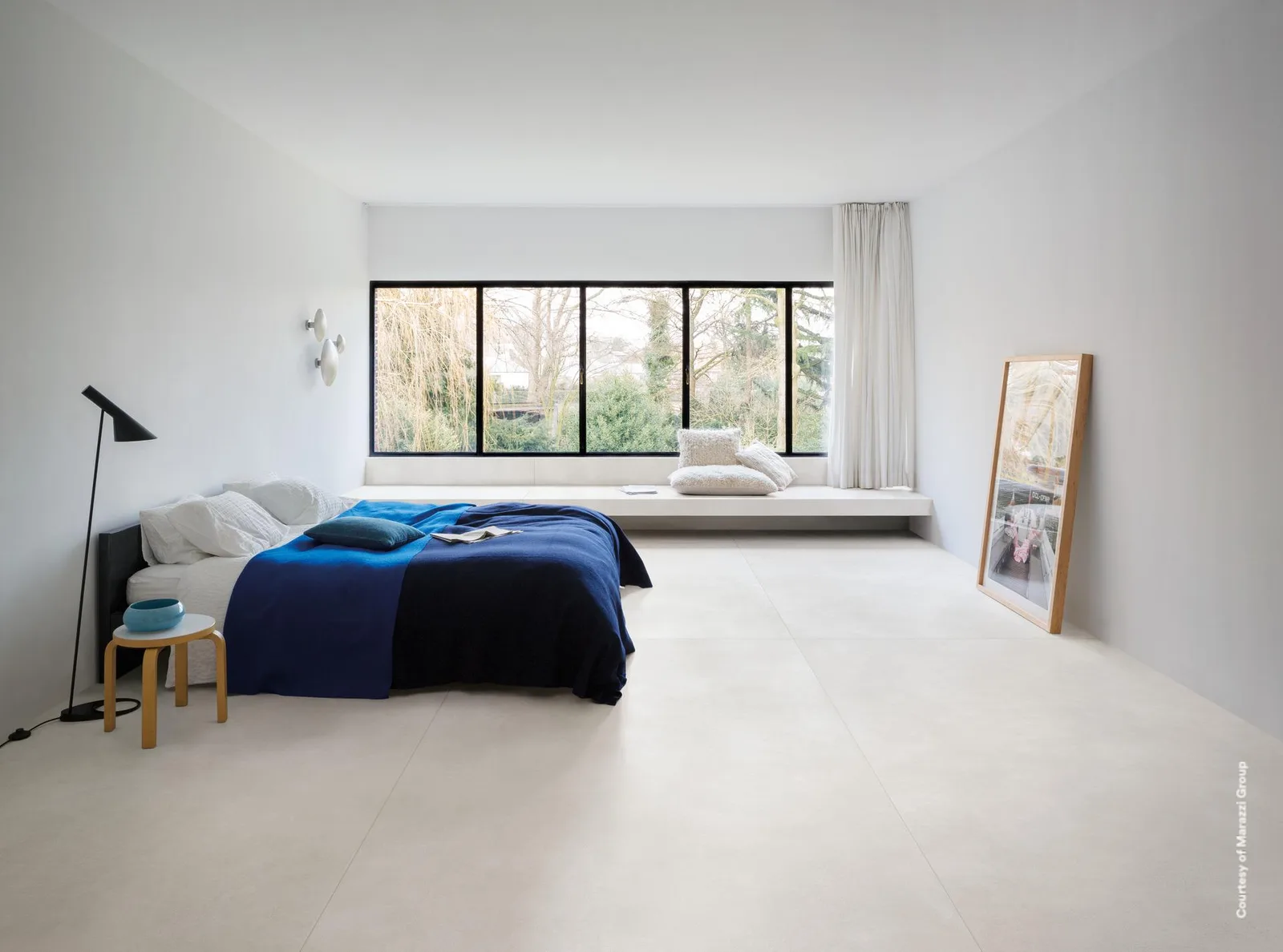 Helles Schlafzimmer mit Bett, blauer Tagesdecke und Fensterband auf großformatigem Boden in Weiß.