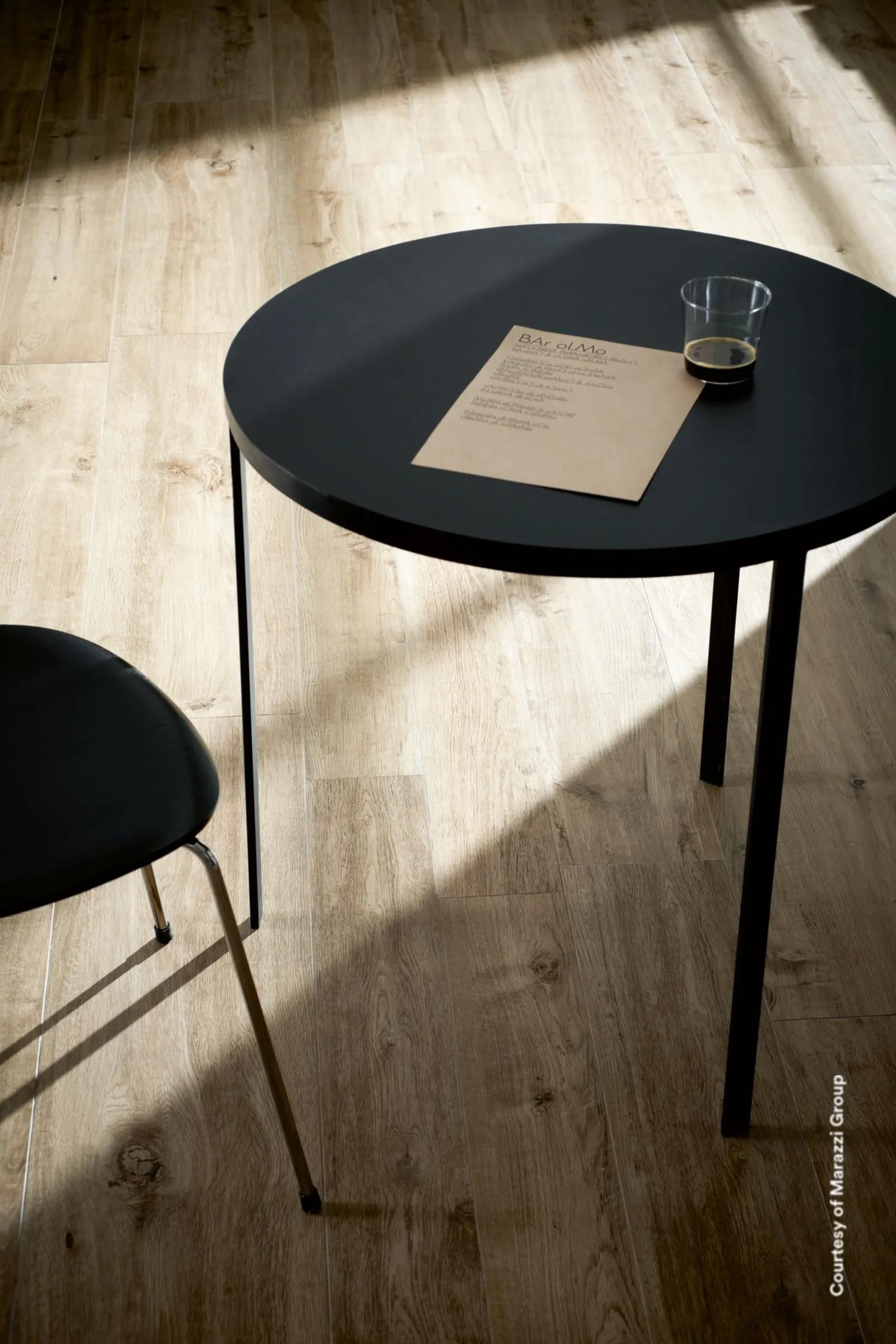 Graubeiger Dielenboden in Holzoptik mit Ästen, darauf ein runder schwarzer Tisch und ein Stuhl im Sonnenlicht.