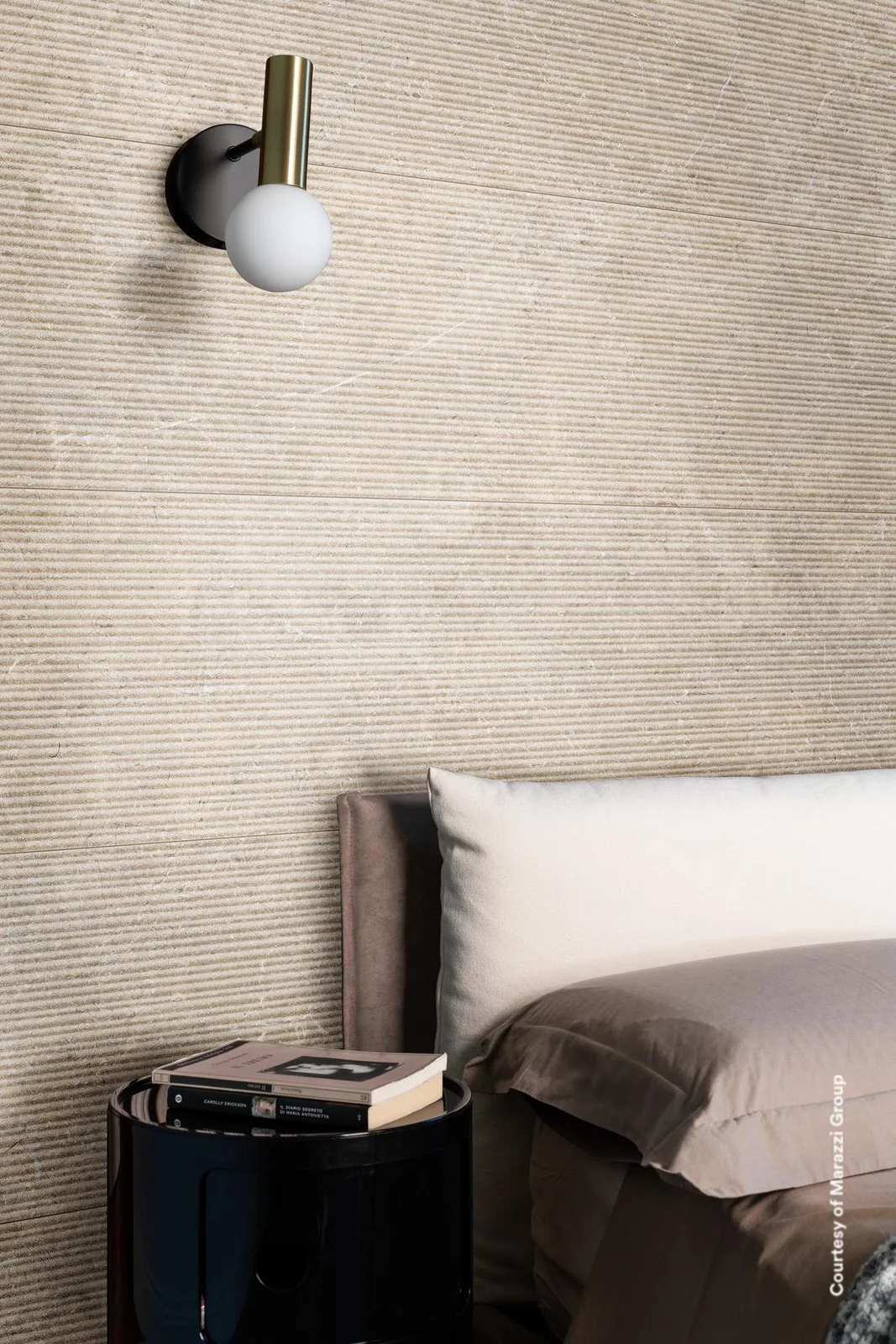 Schlafzimmerwand mit sandbeigen Fliesen mit Rillenrelief hinter dem Bett, dazu eine Wandleuchte und ein Nachttisch.