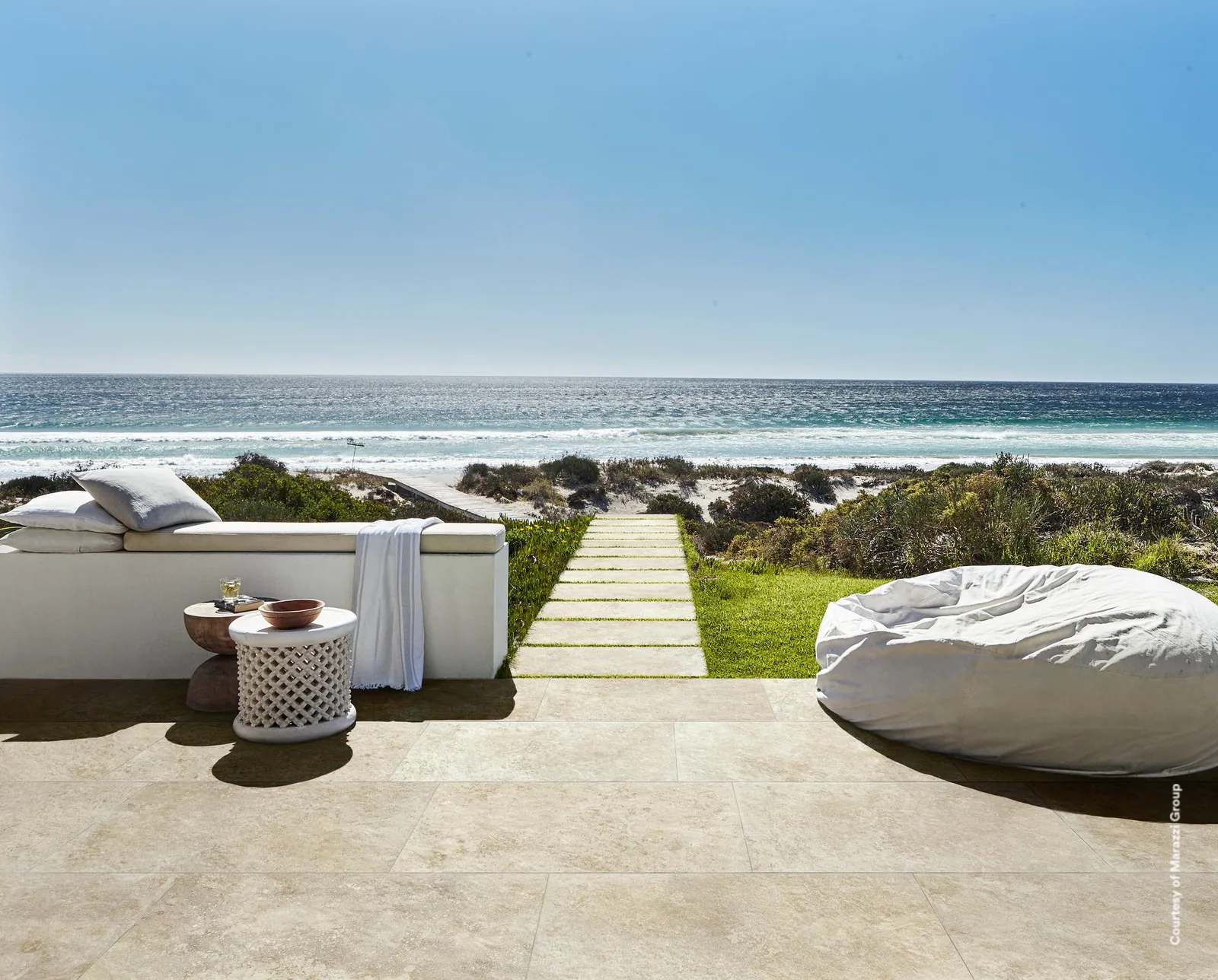 Terrasse am Meer mit hellbeigen Großformatplatten, weißer Sitzbank, Sitzsack und Trittplattenweg durch die Dünen.