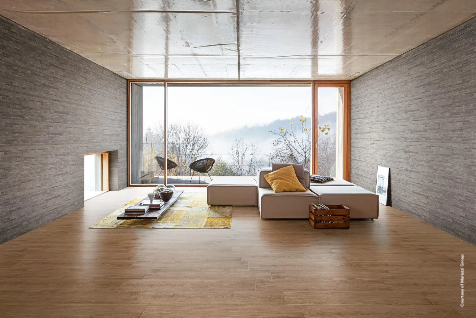 Wohnraum mit großer Fensterfront, weißem Sofa und rötlich-braunem Holzoptik-Boden zwischen grauen Steinwänden.
