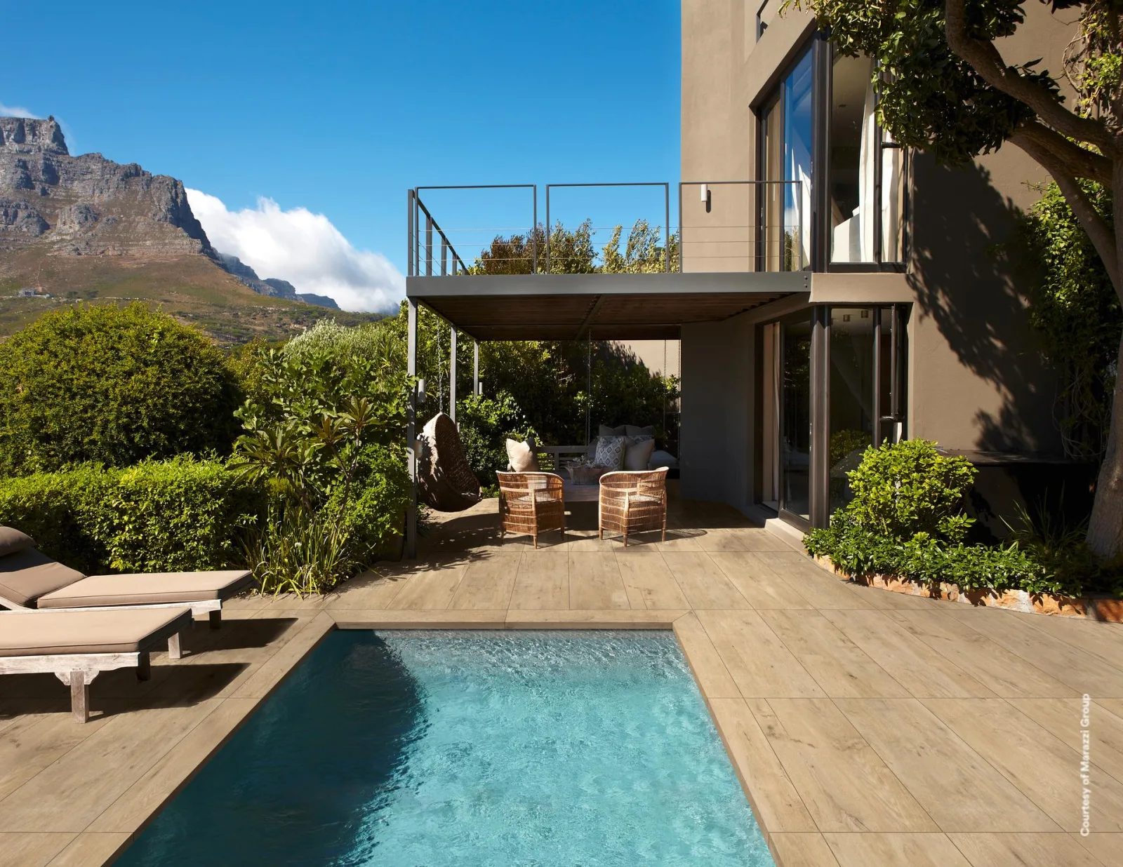 Poolterrasse mit beigefarbenen Holzoptik-Fliesen, Liegen am Beckenrand und Bergkulisse im Hintergrund.